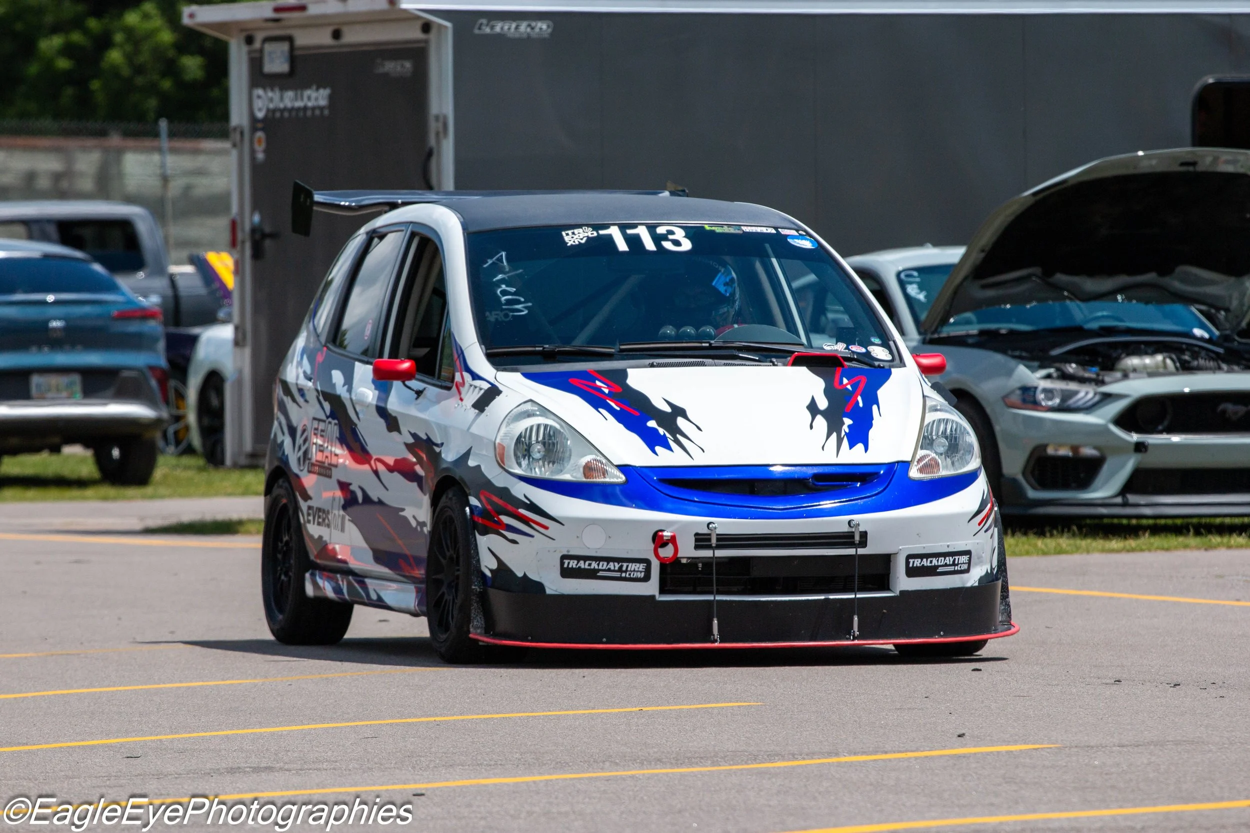 A race car with a white, blue, and red design and the number 113 on the windshield, parked in a lot with other cars and a trailer in the background.