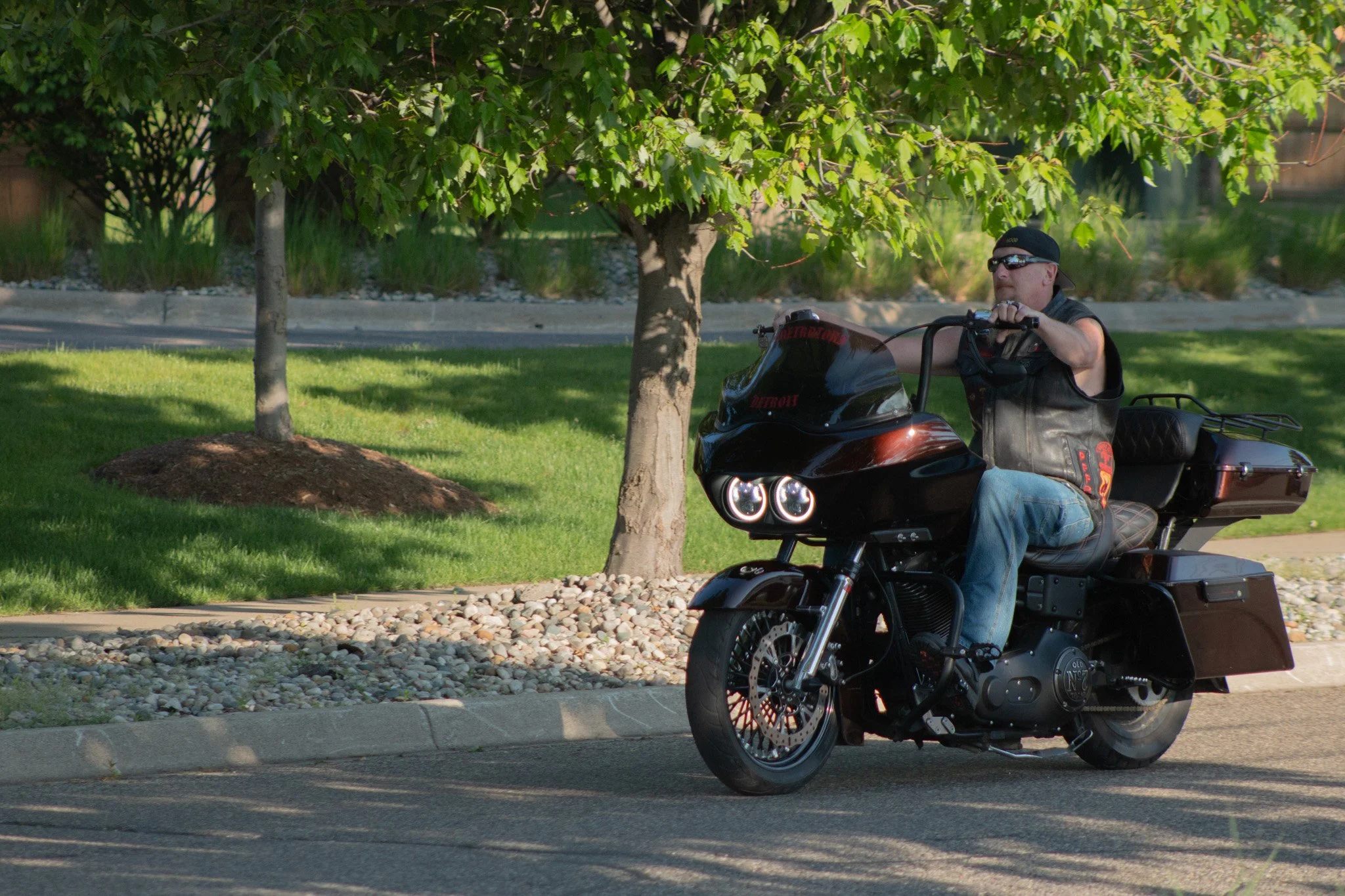 A man riding a black motorcycle in a park with green trees and grass, wearing sunglasses, a sleeveless vest, and a black cap worn backwards.