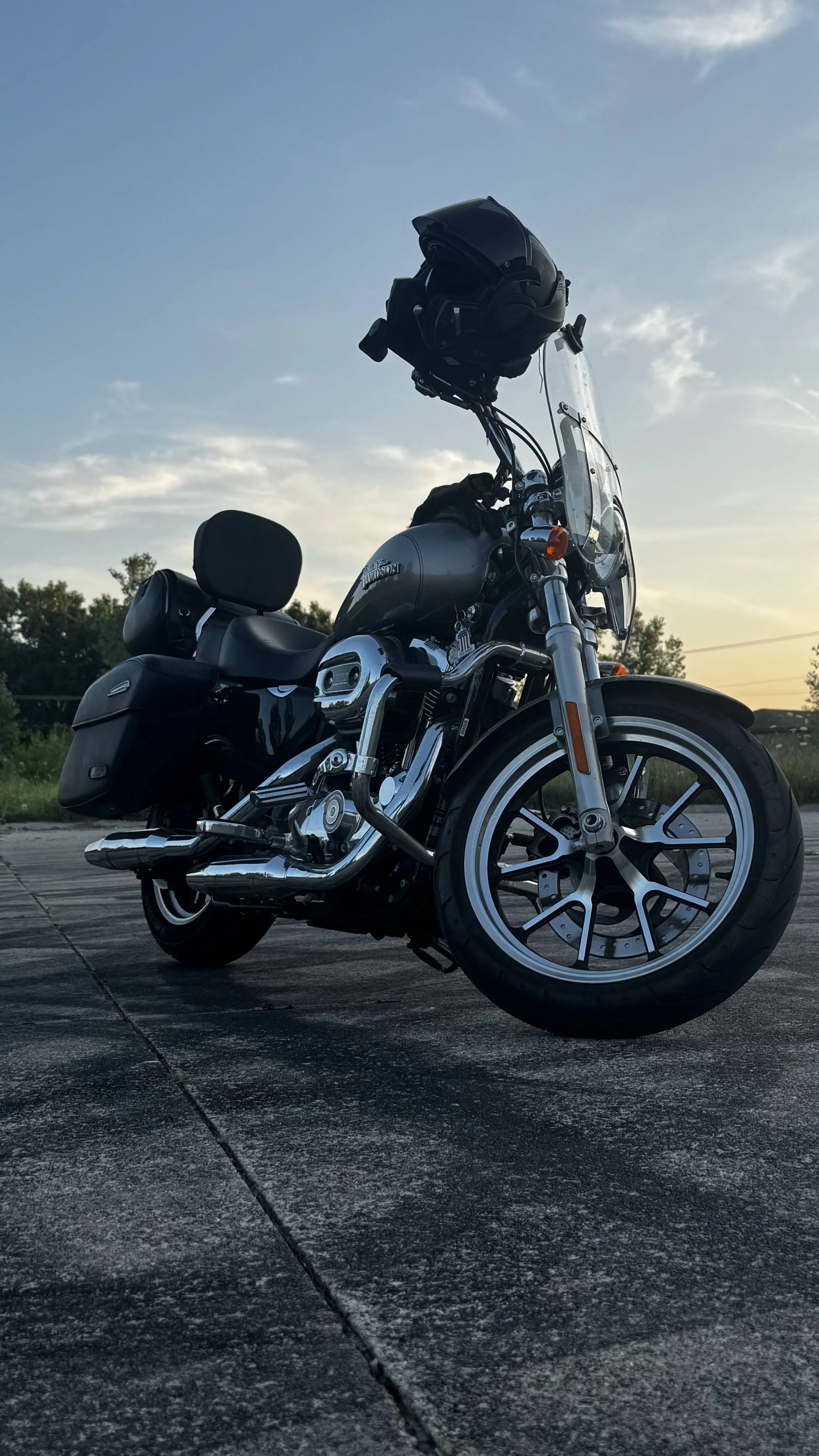 A black motorcycle with saddlebags parked on a paved surface during sunset, with trees and a cloudy sky in the background.