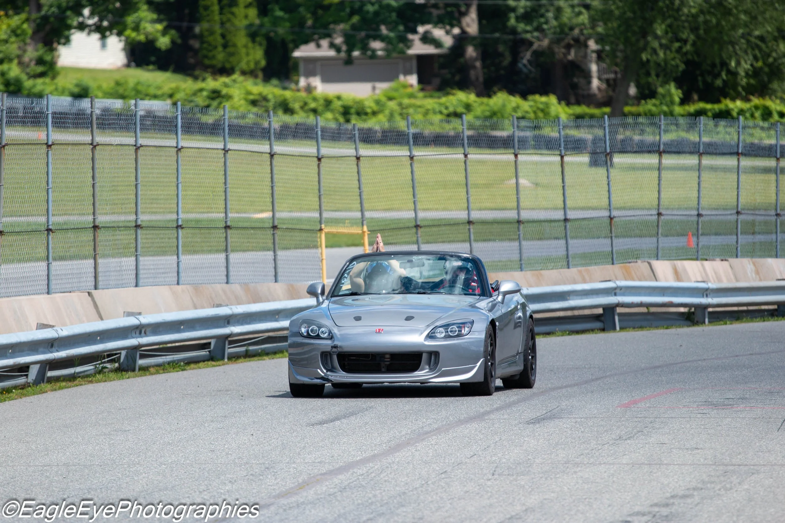 A silver convertible sports car driving on a curved road with a metal guardrail, in a sunny outdoor setting with green trees and fences in the background.