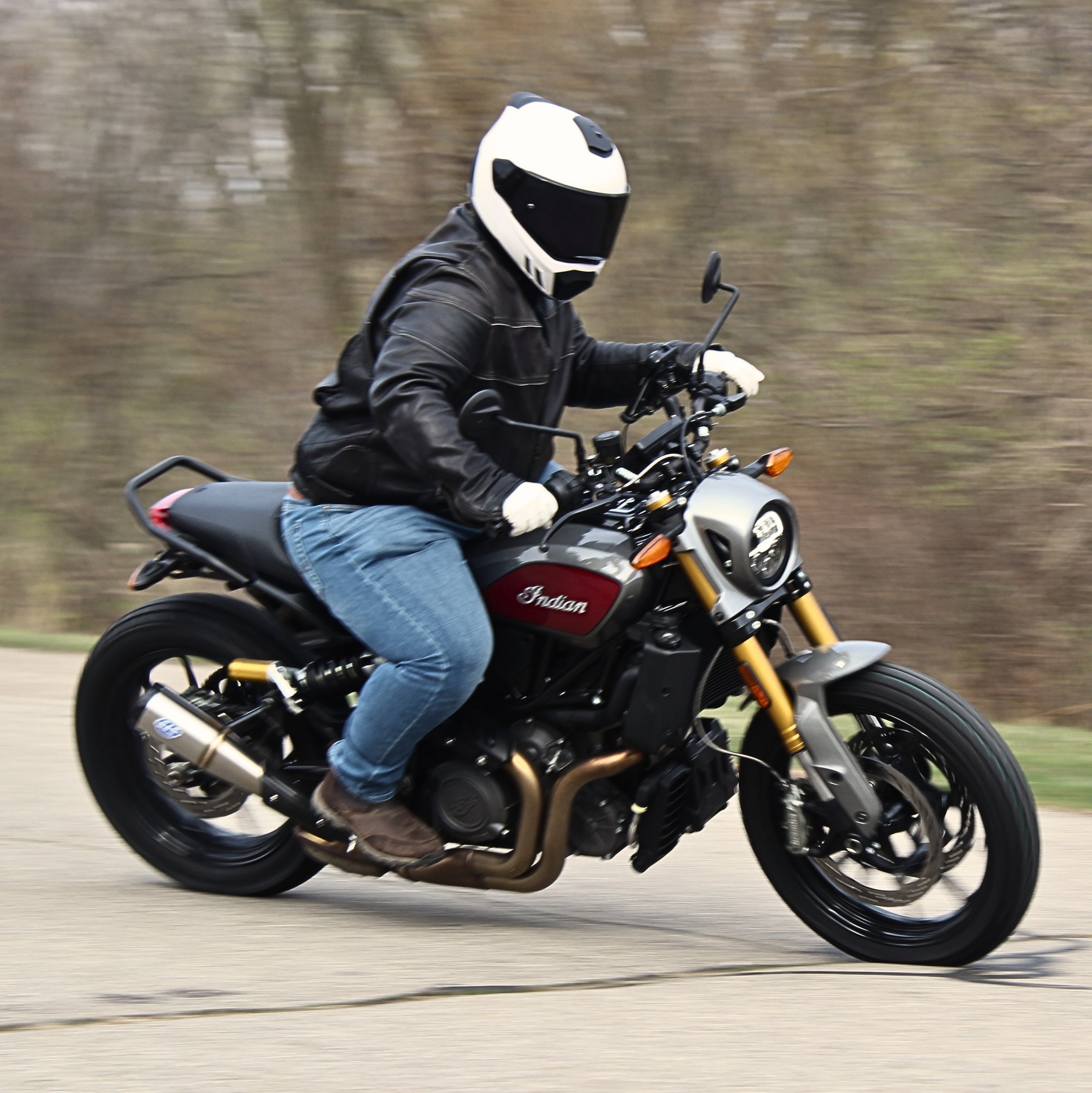Person riding an Indian motorcycle with a white helmet and black leather jacket on a paved road through a wooded area.