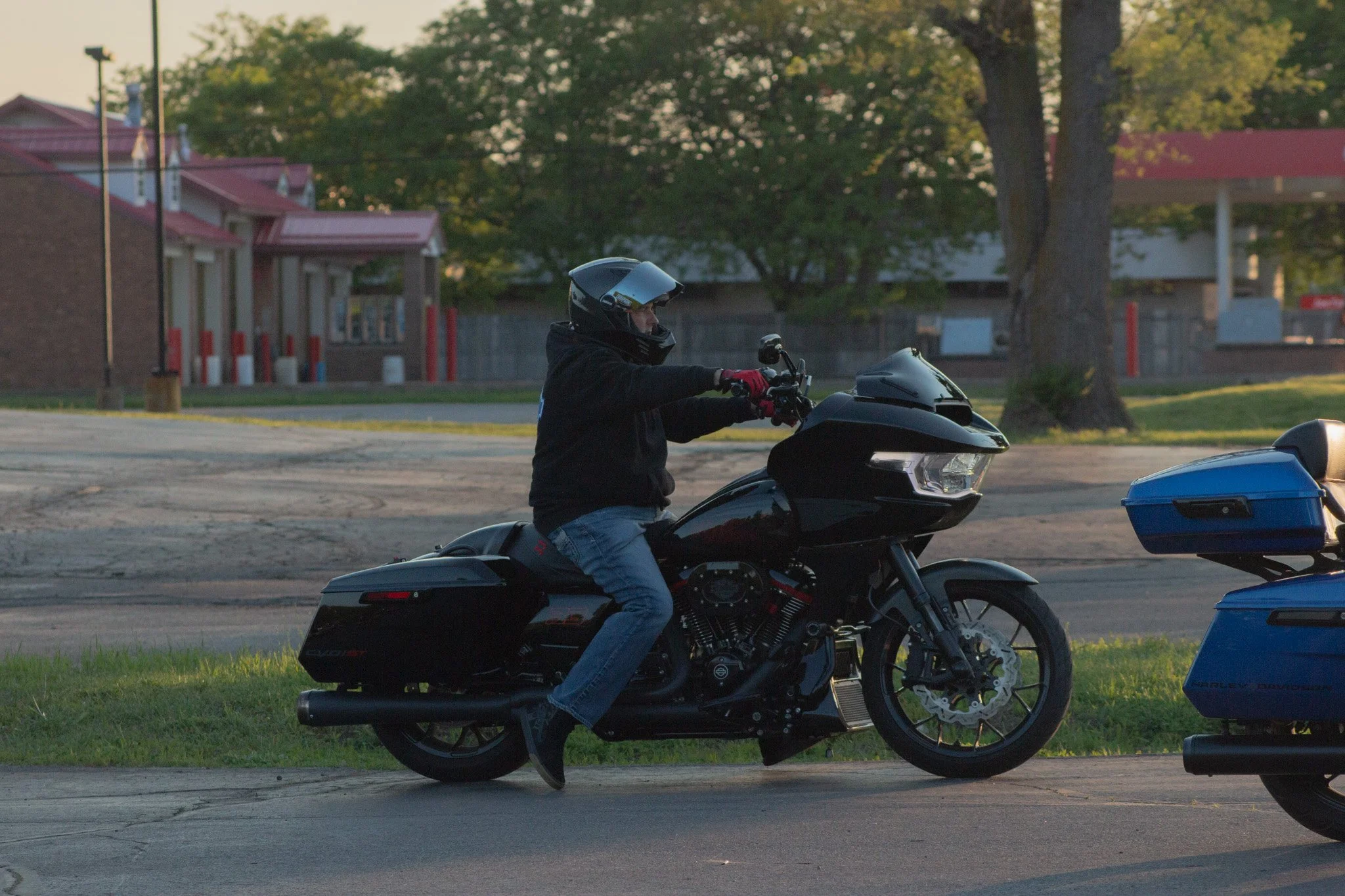 A person riding a black motorcycle with a black helmet and red gloves on a street during daylight. There are trees and buildings in the background.