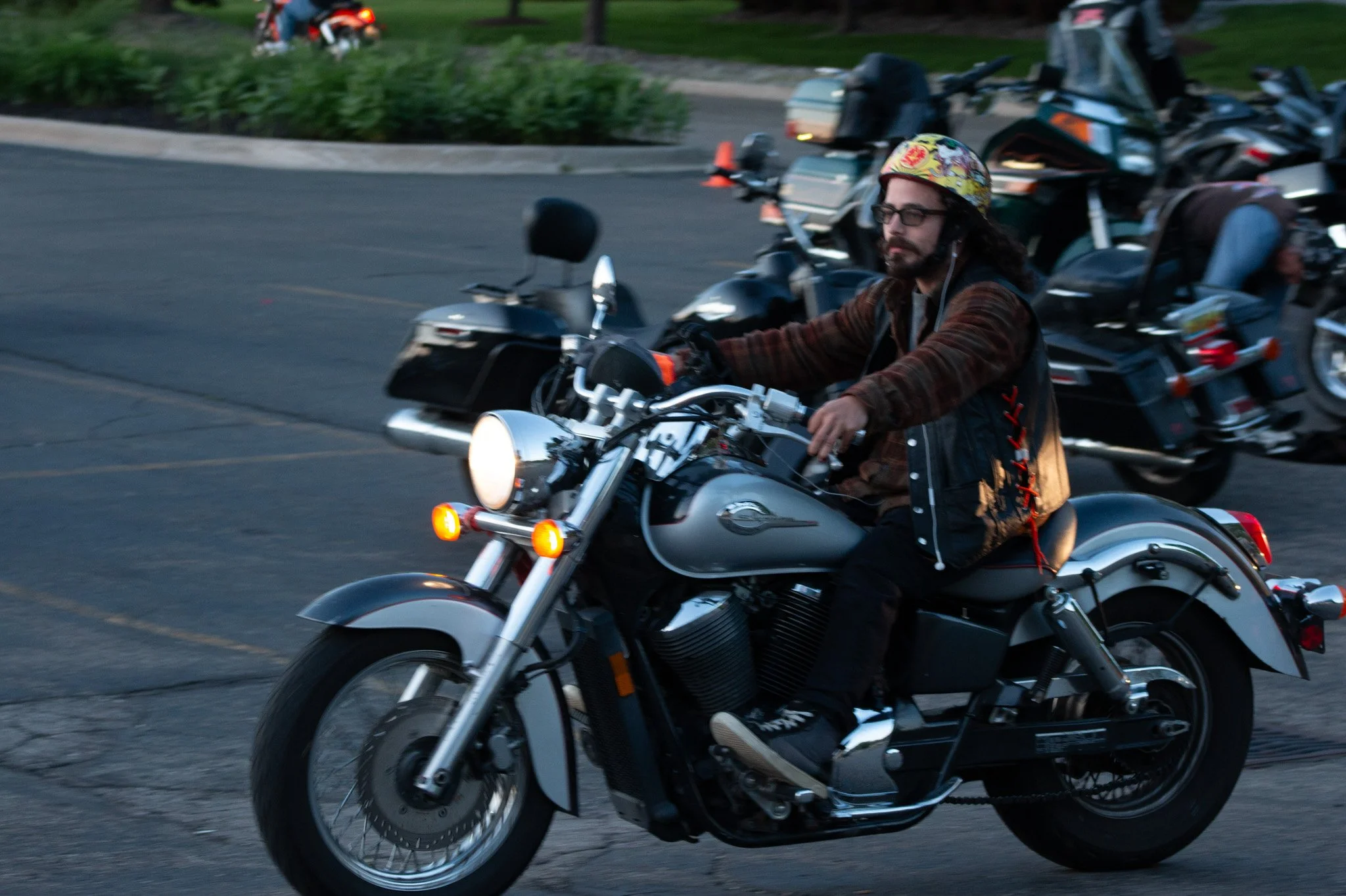 A man with long hair, beard, glasses, wearing a colorful helmet, brown shirt, and vest riding a black and silver motorcycle on a city street, with other motorcycles and greenery in the background.