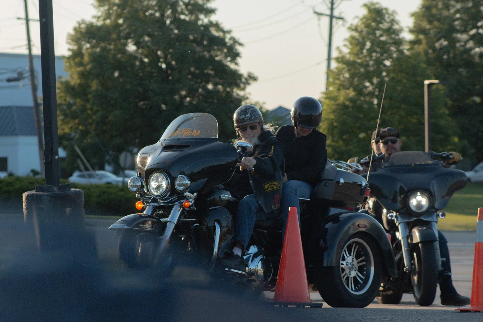 Two people riding a motorcycle past orange safety cones during daylight with trees and power lines in the background.