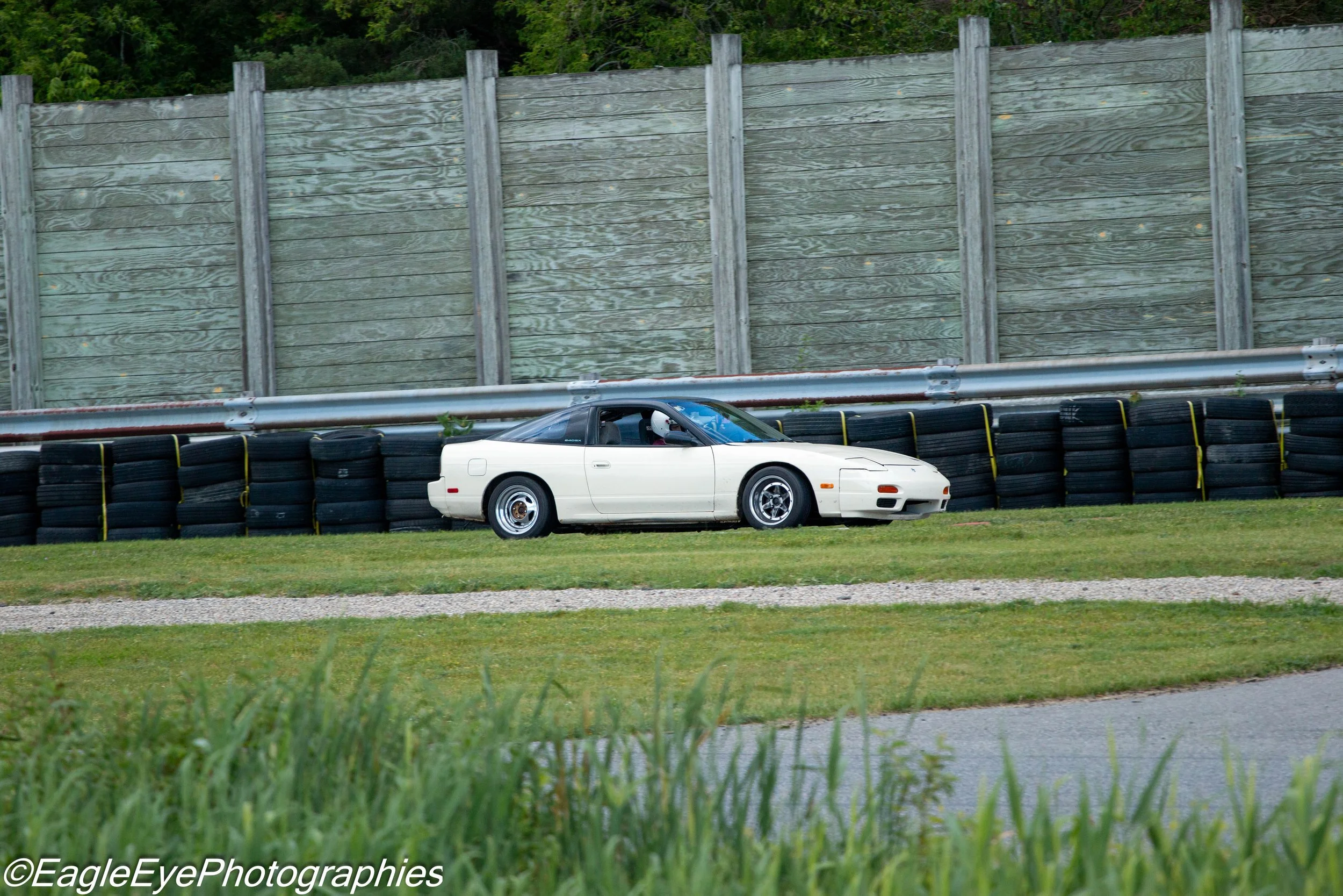 A white sports car driving on a race track with tires stacked along the side and a tall wooden barrier, surrounded by green trees.