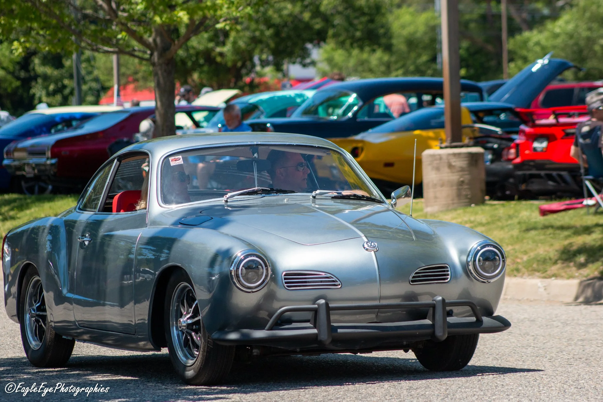 A silver vintage car with round headlights and a front bumper guard, parked outdoors at a car show, with other classic cars and trees in the background.