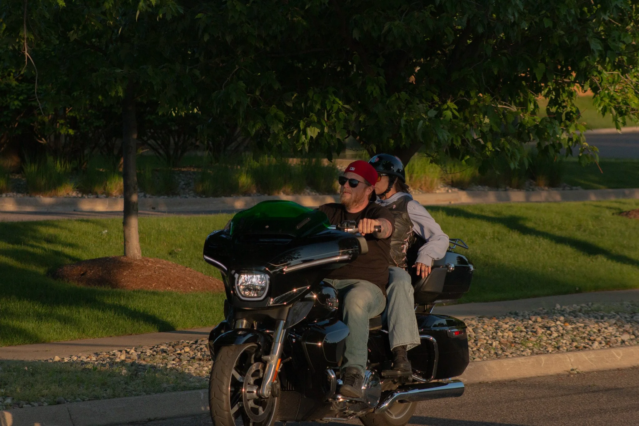 Two people riding a black motorcycle along a tree-lined street during daytime. The rider is wearing sunglasses, a black helmet, and a red cap. The passenger is wearing a black helmet and has long hair. There is greenery and a sidewalk with rocks in t