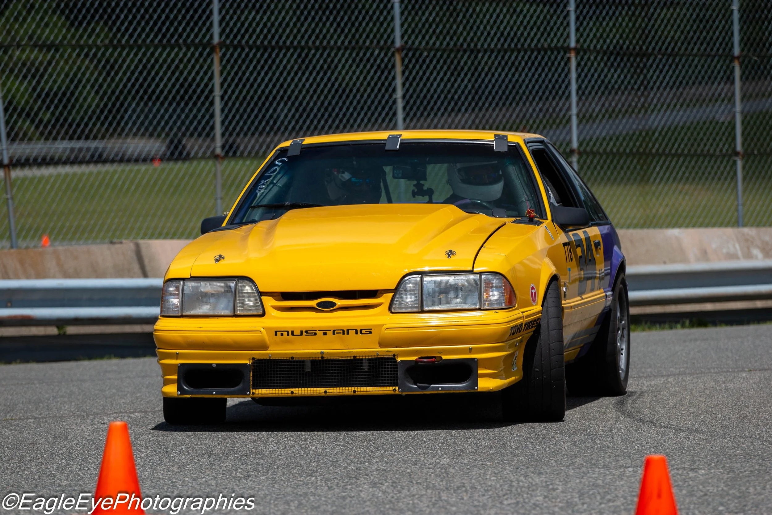 Yellow Ford Mustang race car on a track, with orange cones and a chain-link fence in the background.