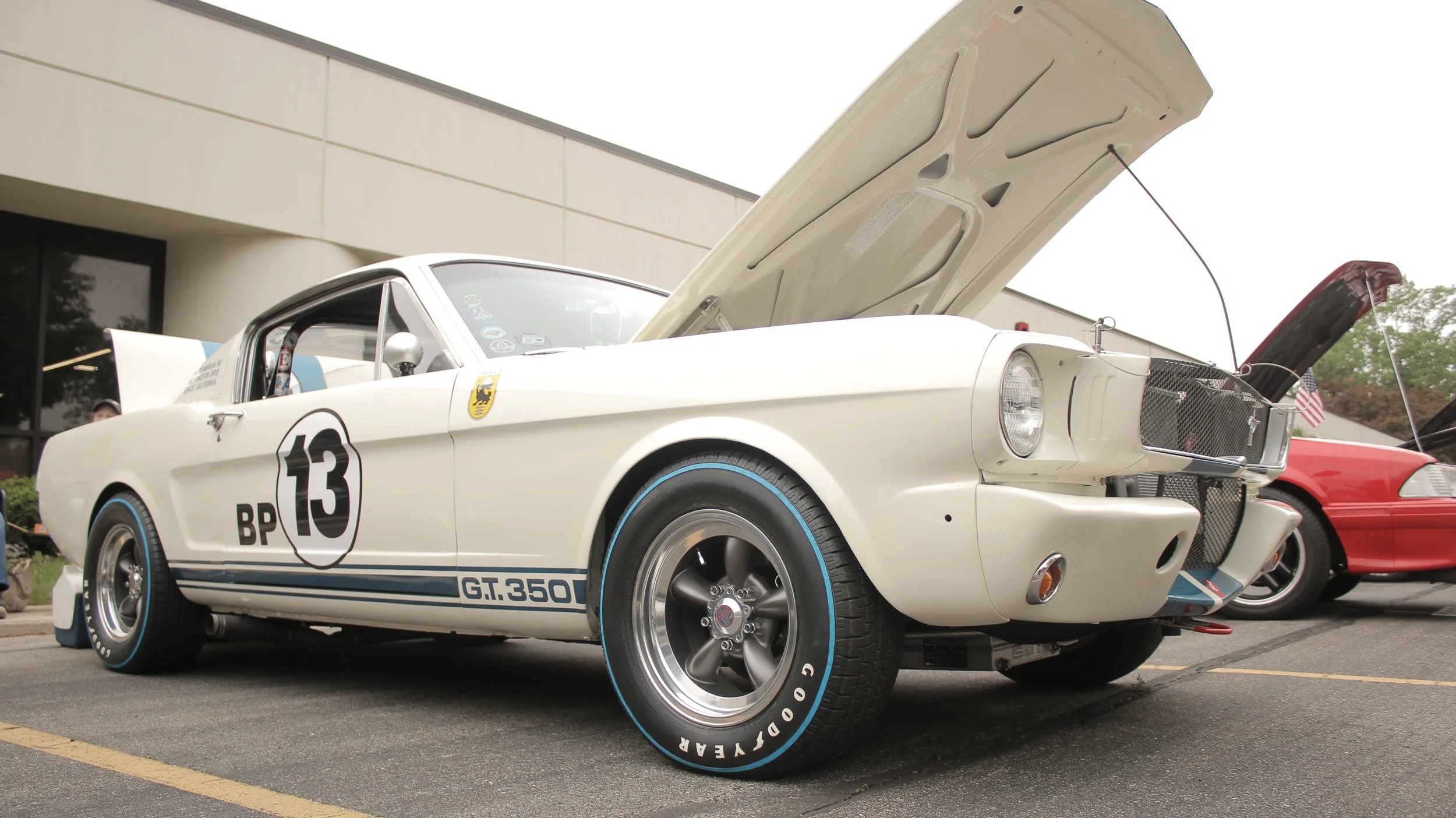 A vintage white race car with open hood displaying engine, parked outdoors near a modern building, with a red car partially visible beside it.
