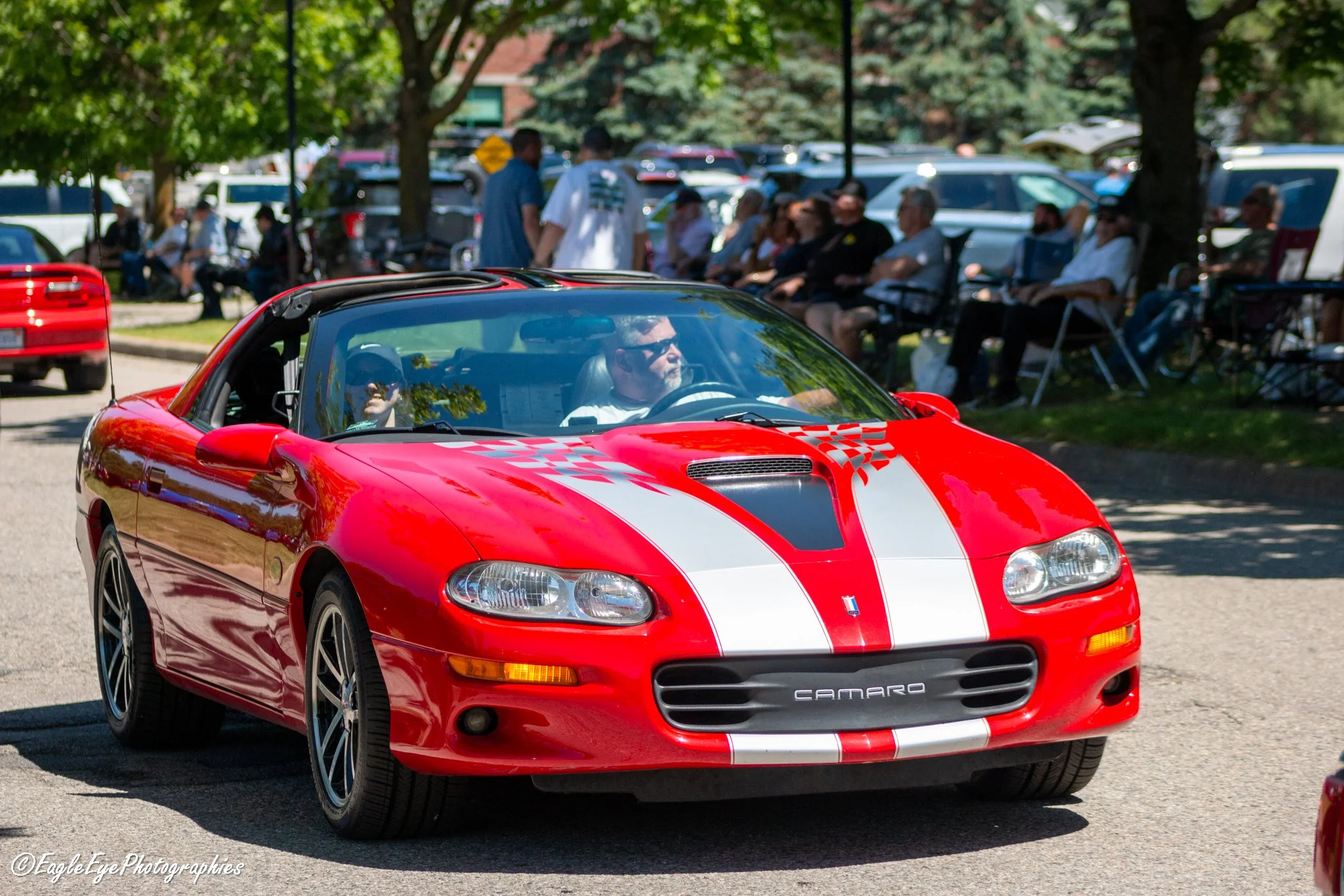 A red Chevrolet Camaro sports car with white racing stripes driving past a crowd of people sitting on chairs under trees at an outdoor event.