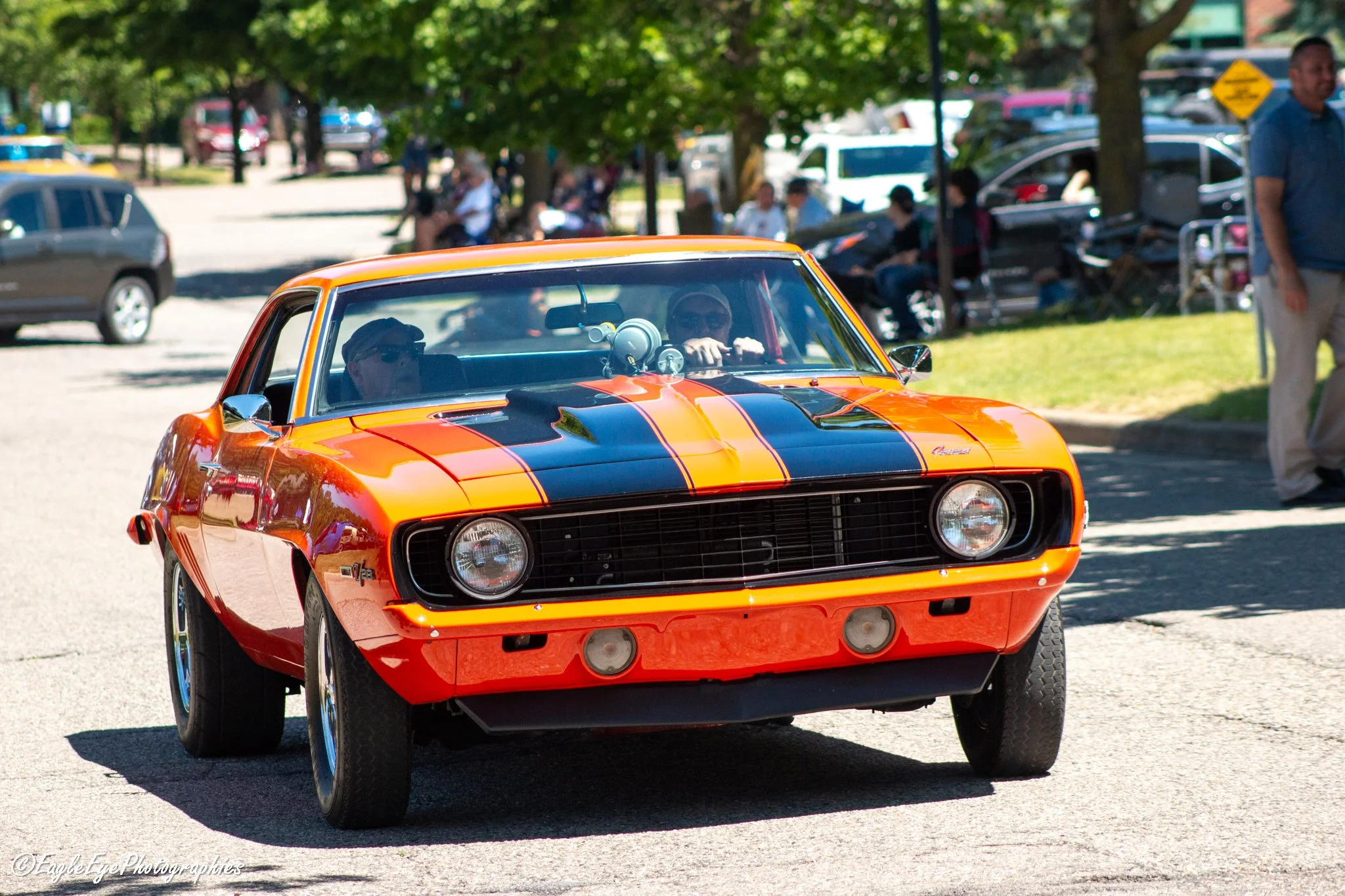 A vintage orange car with black racing stripes driving on a street with people sitting on benches and trees lining the background.