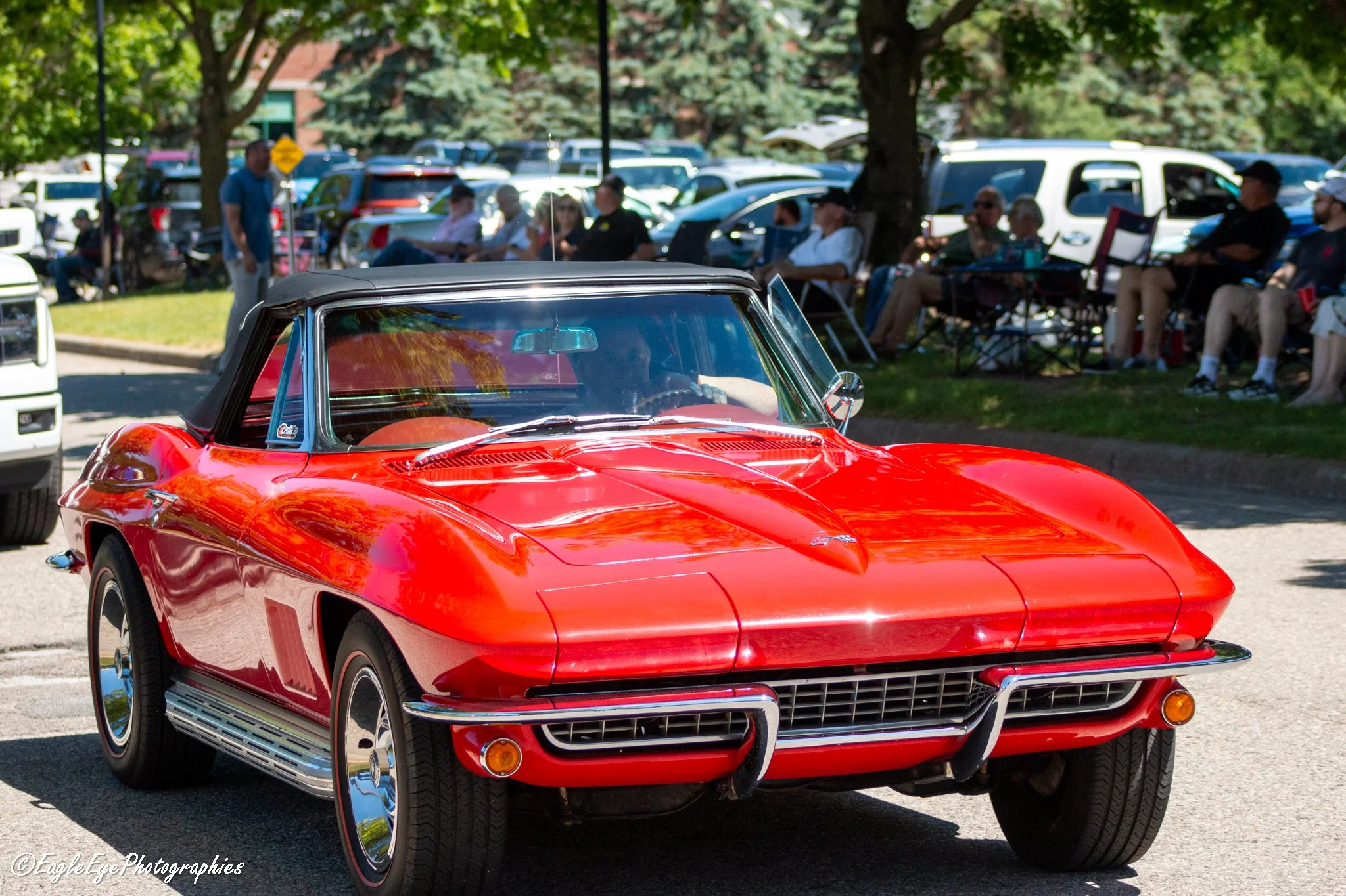 A red vintage convertible car parked in a parking lot during a car show. In the background, people sitting under trees and a row of parked cars can be seen.