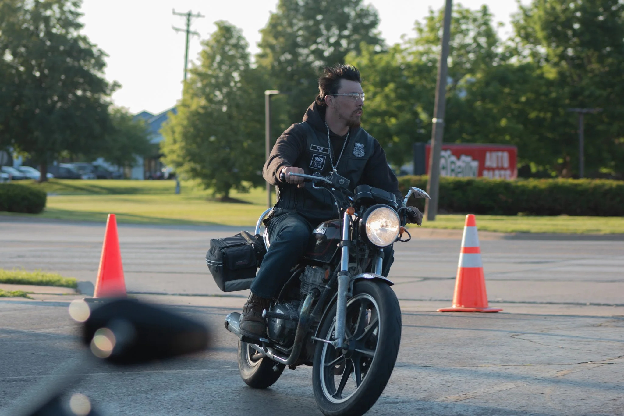 A man with dark hair, glasses, and a beard riding a motorcycle through a parking lot, maneuvering around orange traffic cones, with trees and a building in the background during daylight.