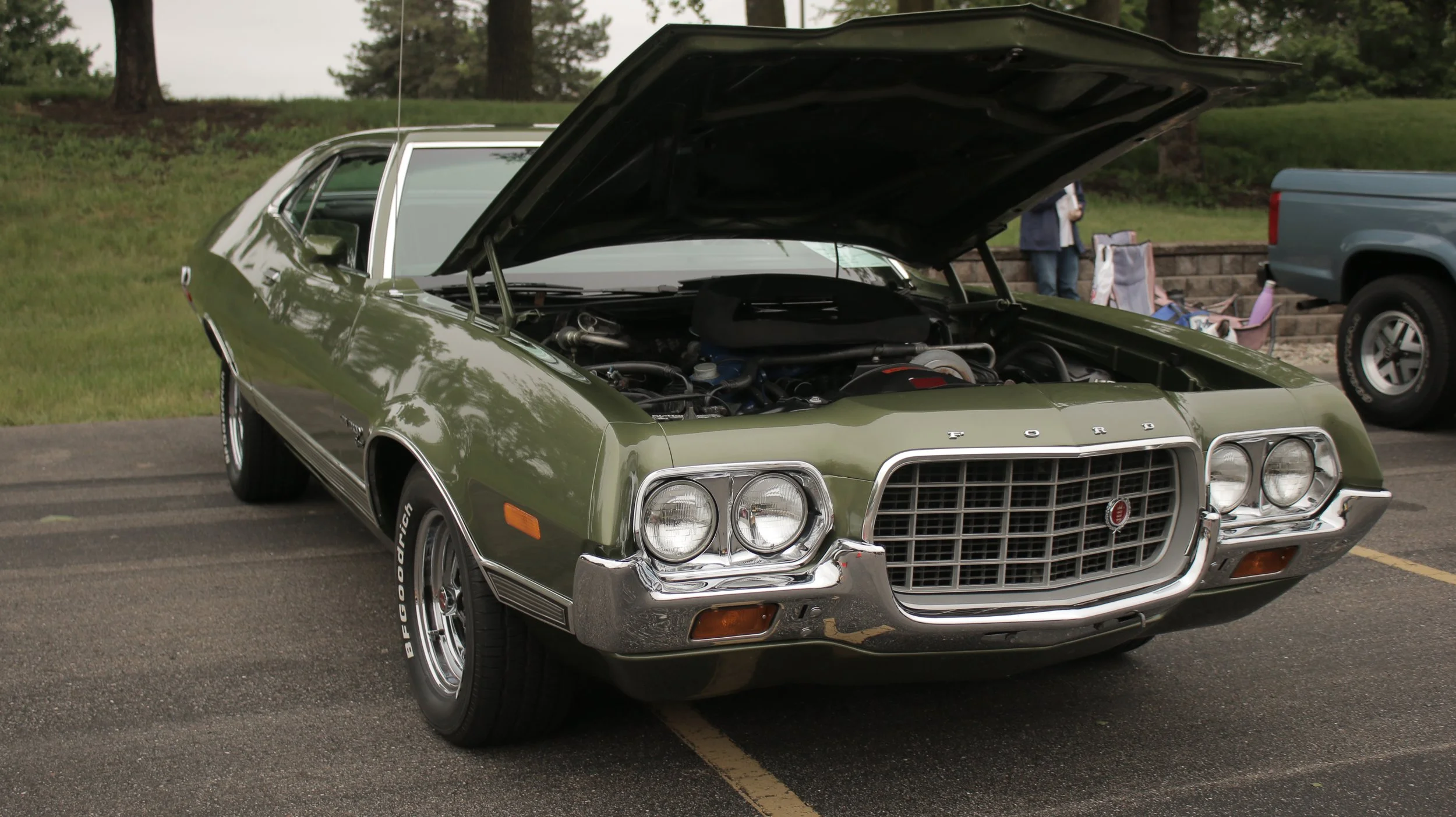 A green vintage Ford car with its hood open, parked on an asphalt lot.