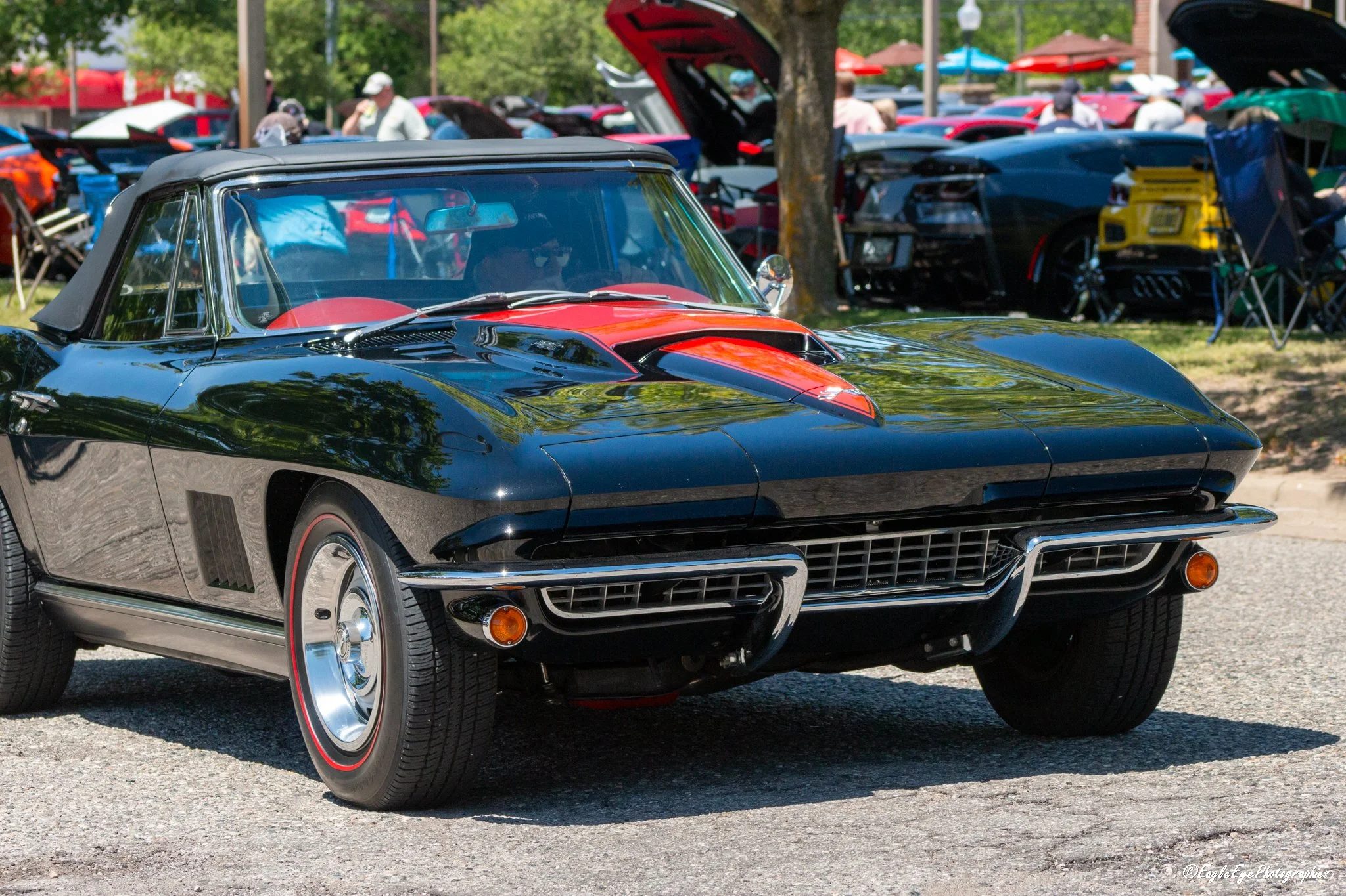 A black classic Corvette with a red stripe and red interior is parked at a car show. In the background, several other cars and people are visible, with some cars with open hoods and tents, under a tree on a sunny day.