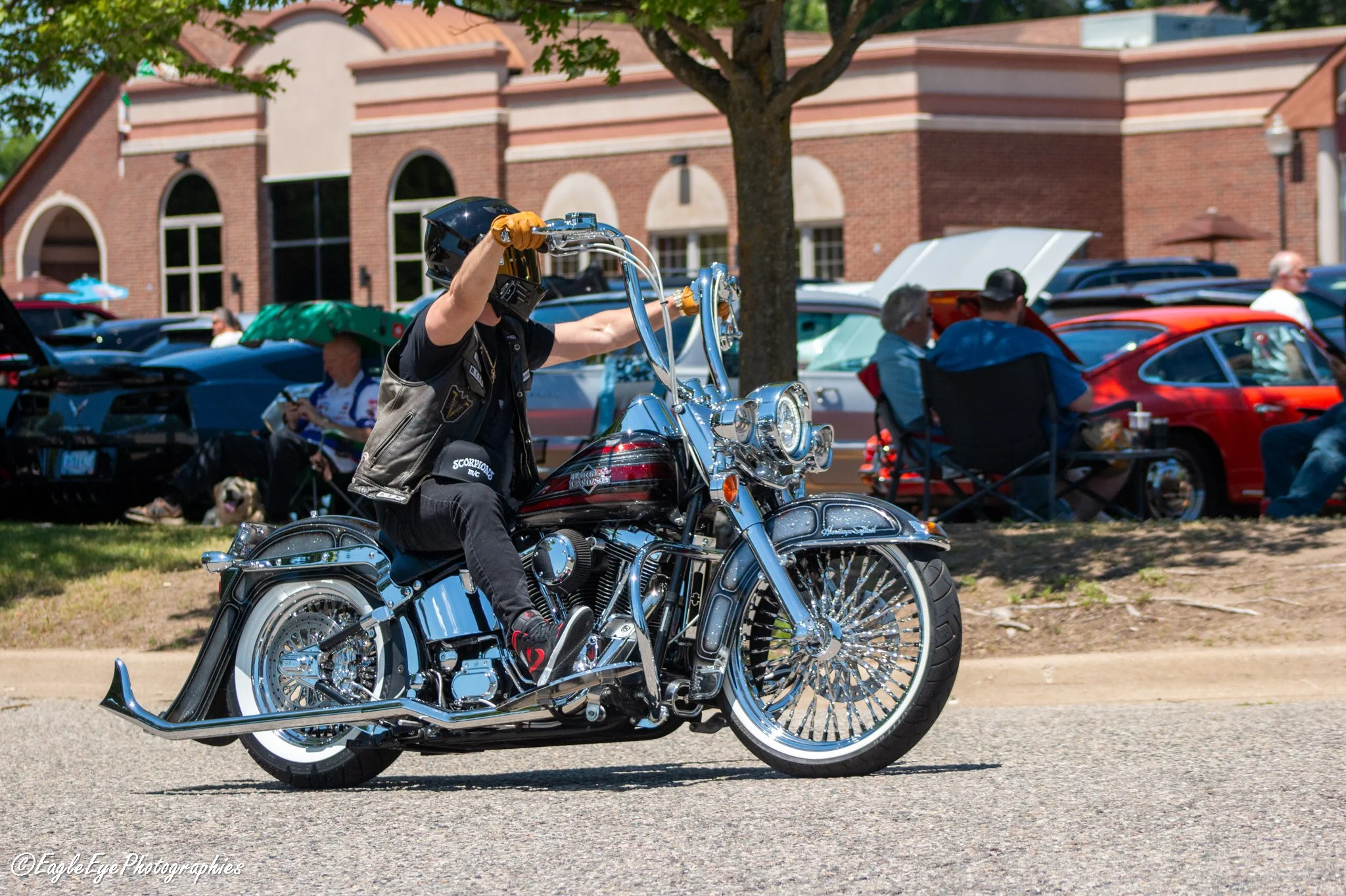 A person riding a shiny black motorcycle with chrome details on a street during a sunny day. They are wearing a helmet, a sleeveless vest, and gloves. Several cars are parked on the side of the street, and a building with arched windows and brick wal