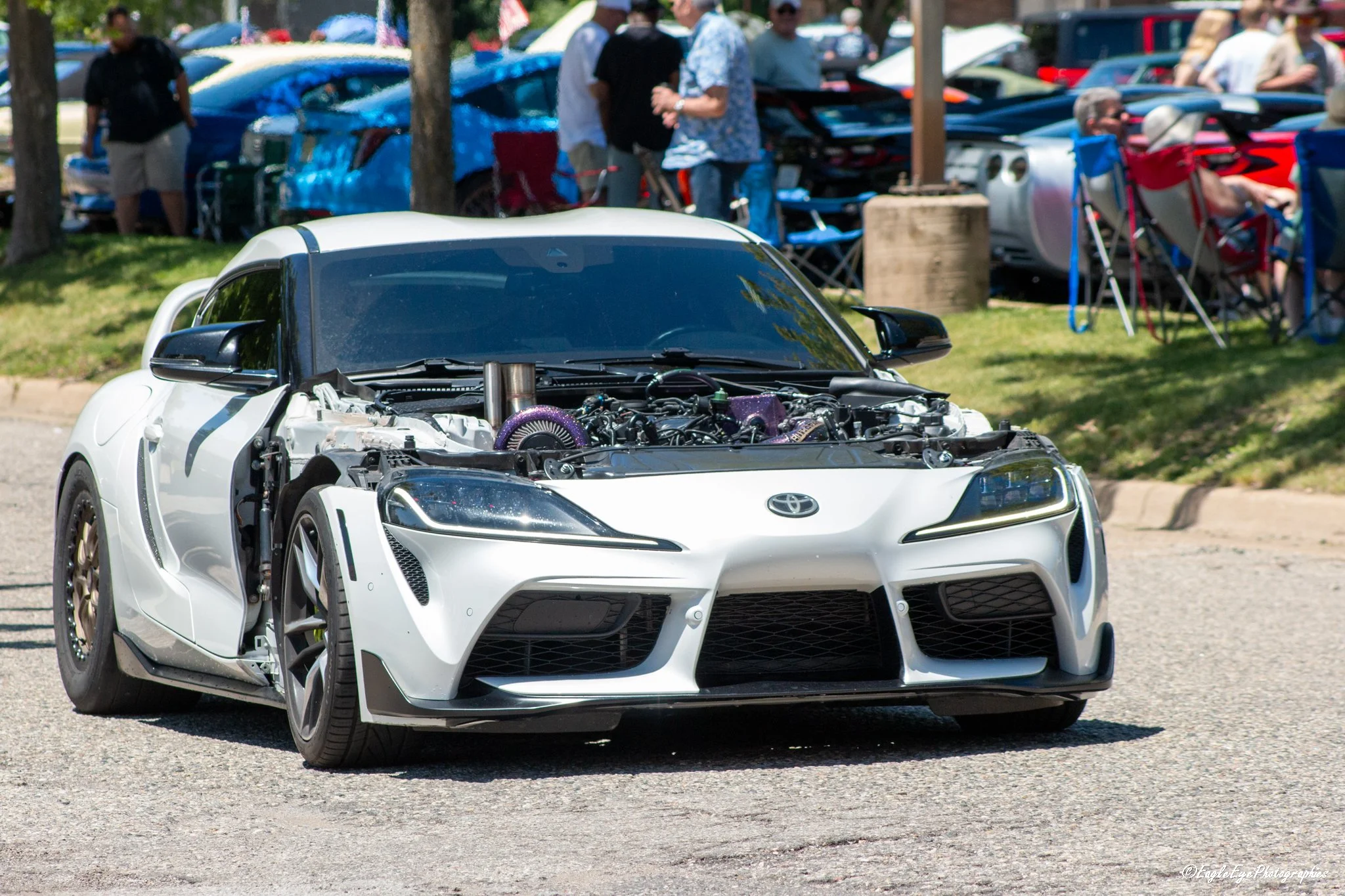 A white sports car with a partially open hood displaying the engine, parked at an outdoor car show with people in the background.