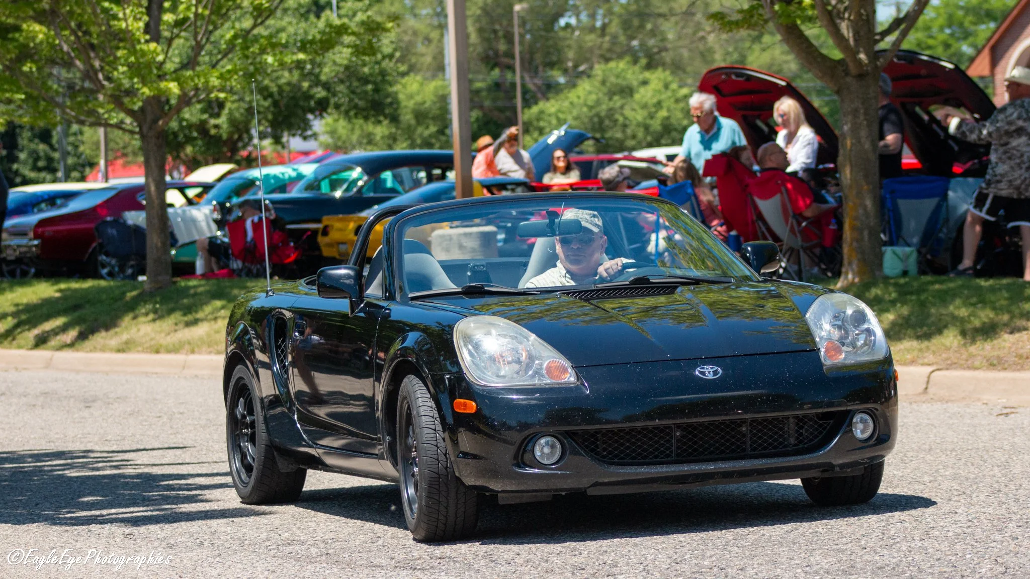 A black convertible sports car driving on a street with a crowd of people under the shade of trees in the background.
