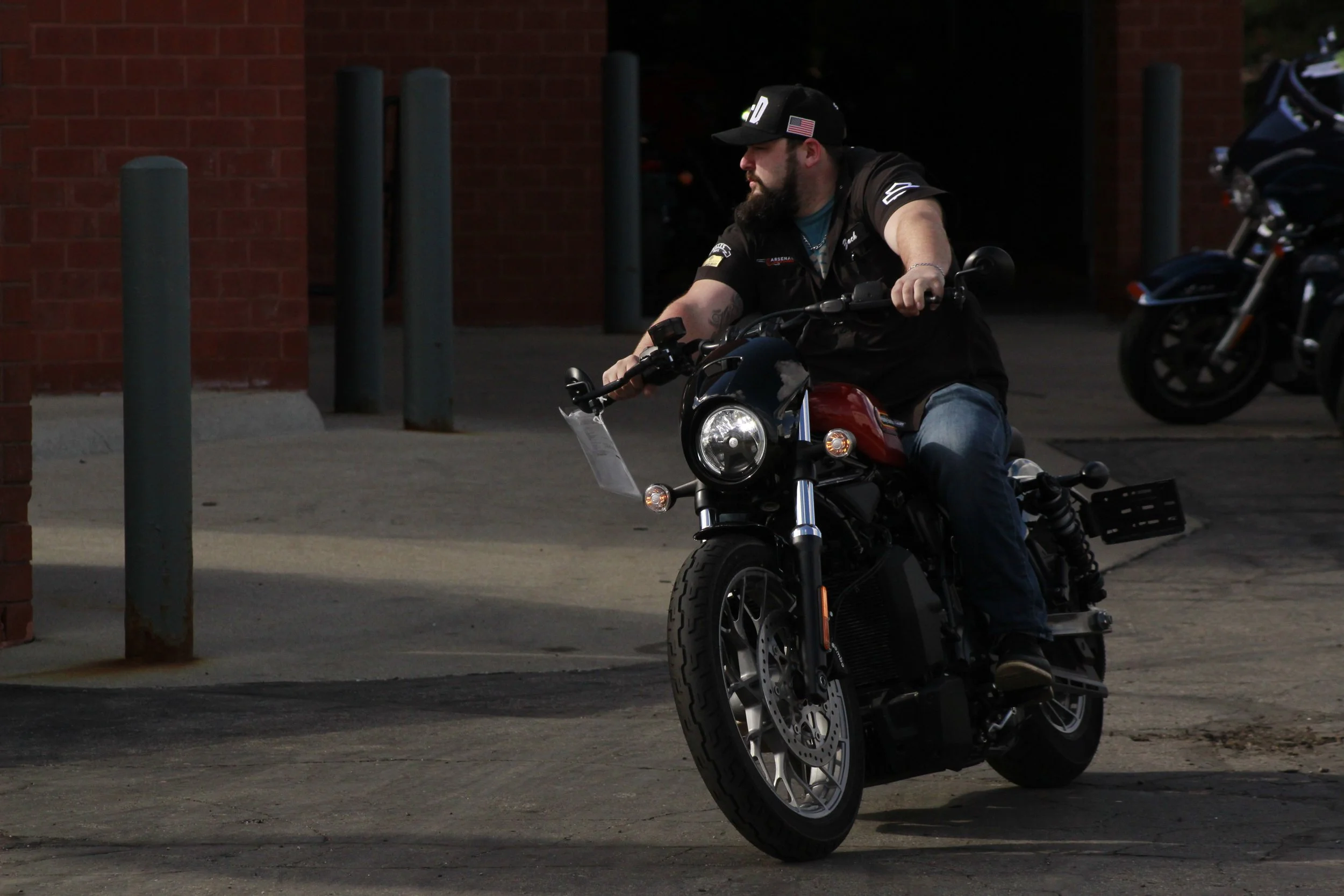 A man with a beard and tattoos riding a black motorcycle on a city street, nearby a brick building with metal bollards.