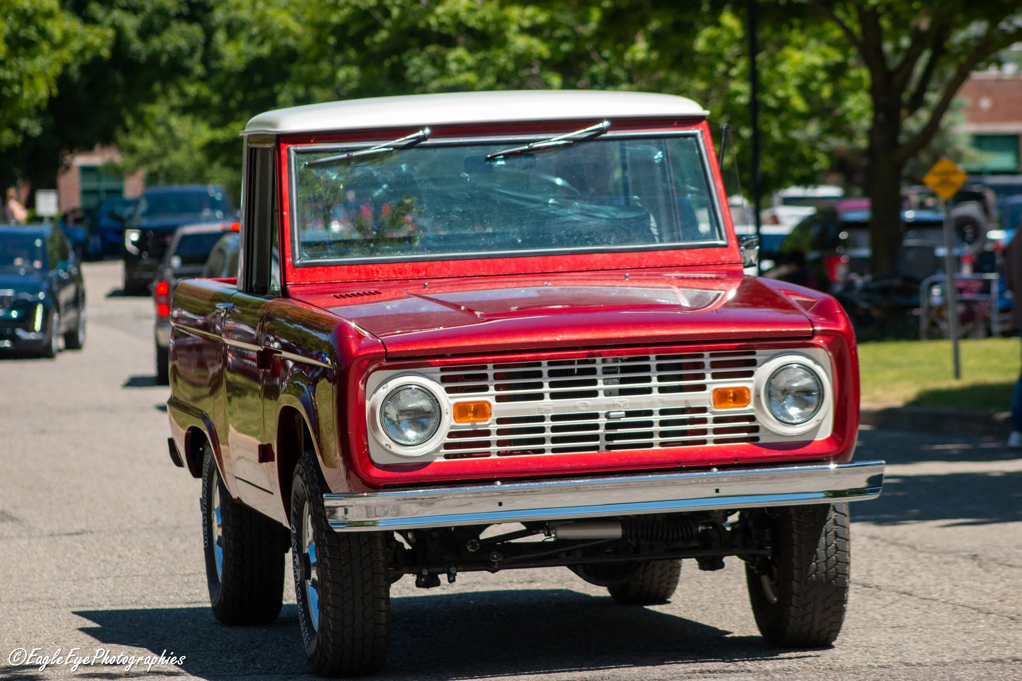 A red vintage Ford Bronco SUV with a white roof parked on a street, with trees and other vehicles in the background.