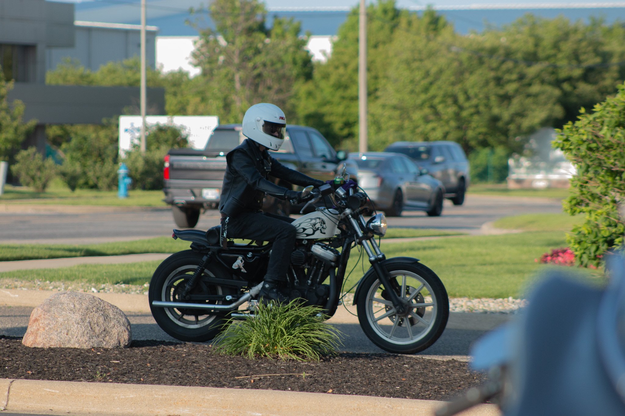 A person riding a motorcycle wearing a white helmet and black clothing on a city street with parked cars, trees, and buildings in the background.