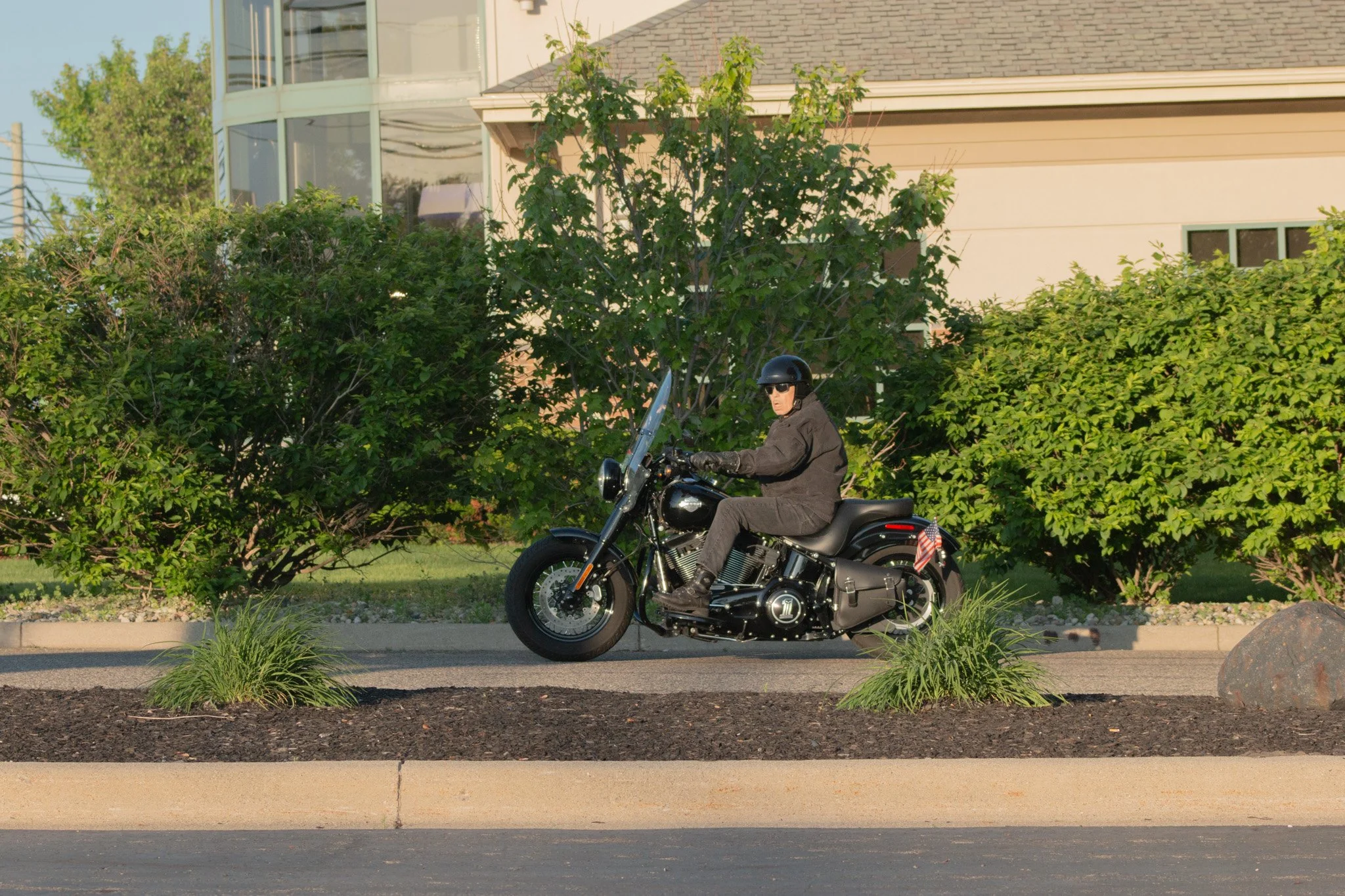 A person wearing a black helmet and sunglasses riding a black motorcycle on a street in front of bushes and suburban houses.