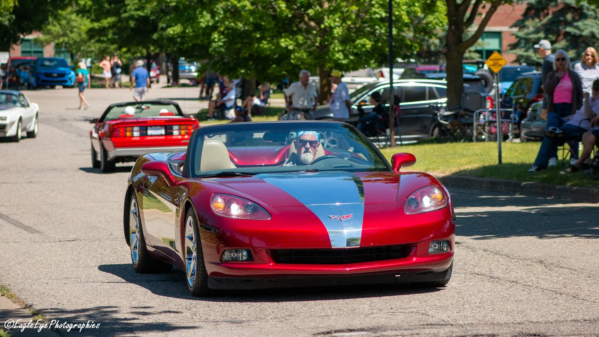 A red Chevrolet Corvette convertible with a silver racing stripe being driven through a crowded outdoor event on a sunny day, surrounded by people sitting and walking under green trees.