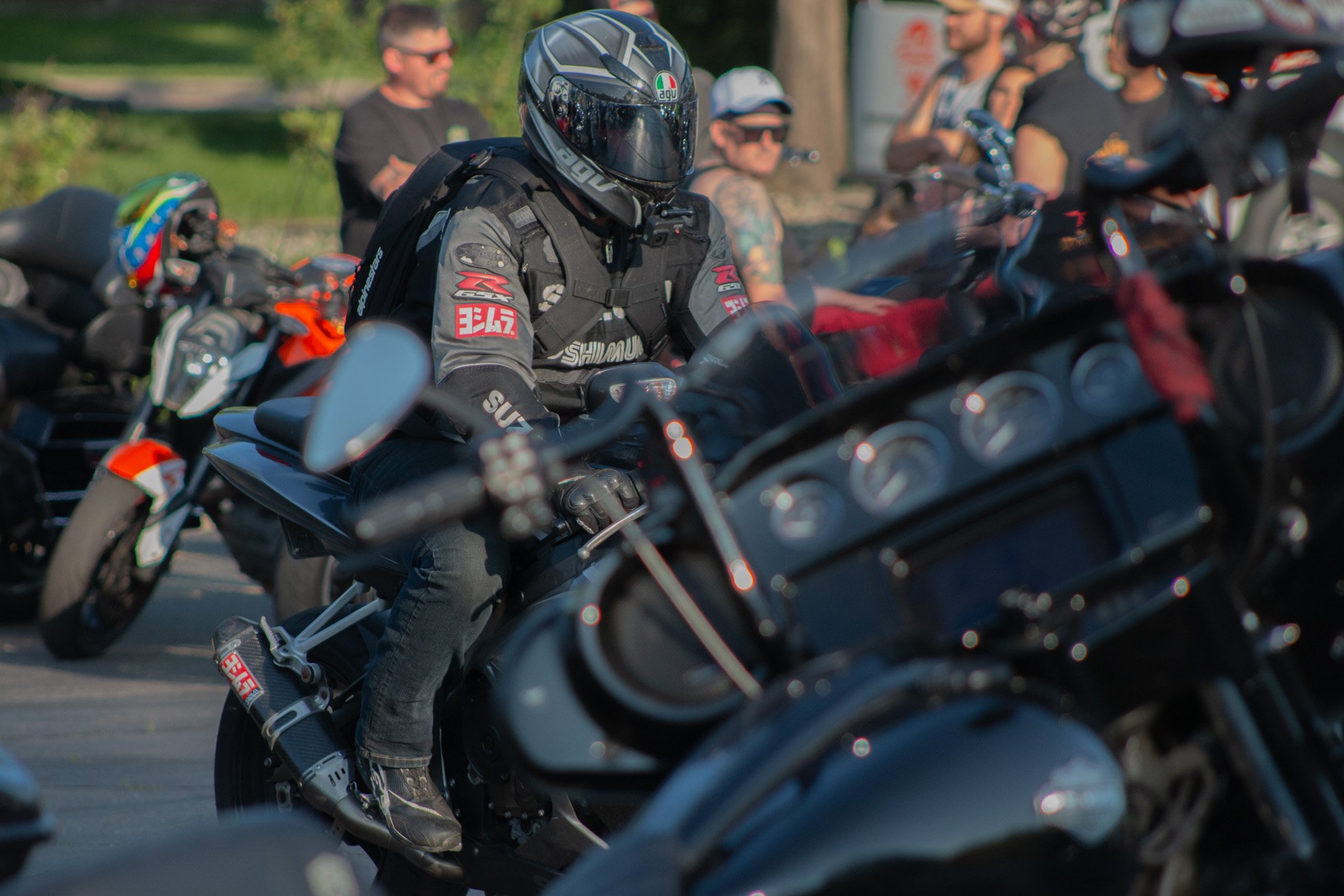 Motorcyclist wearing a black helmet, gray riding jacket, and black gloves sitting on a motorcycle surrounded by other riders and motorcycles in an outdoor setting.