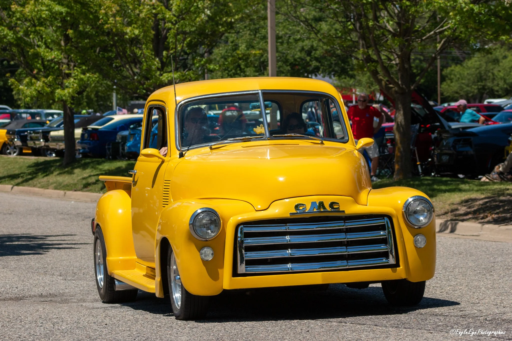 A bright yellow vintage GMC truck driving on a sunny day with trees and parked cars in the background at an outdoor event.