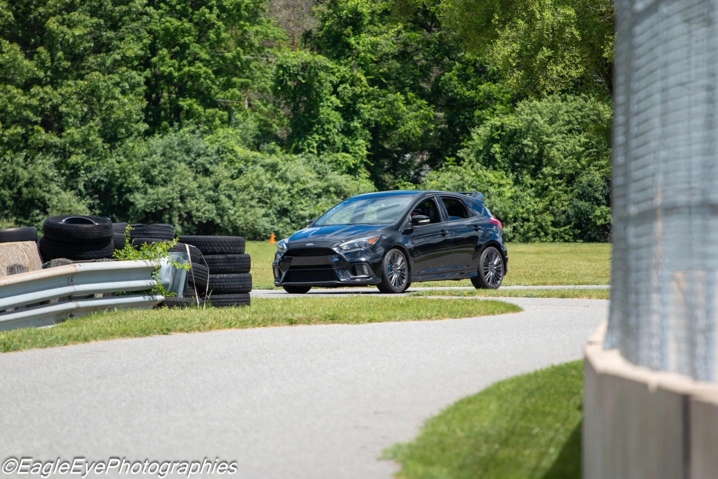 A black Ford Focus parked on a grassy area next to a chain-link fence and a stack of tires, with trees in the background.