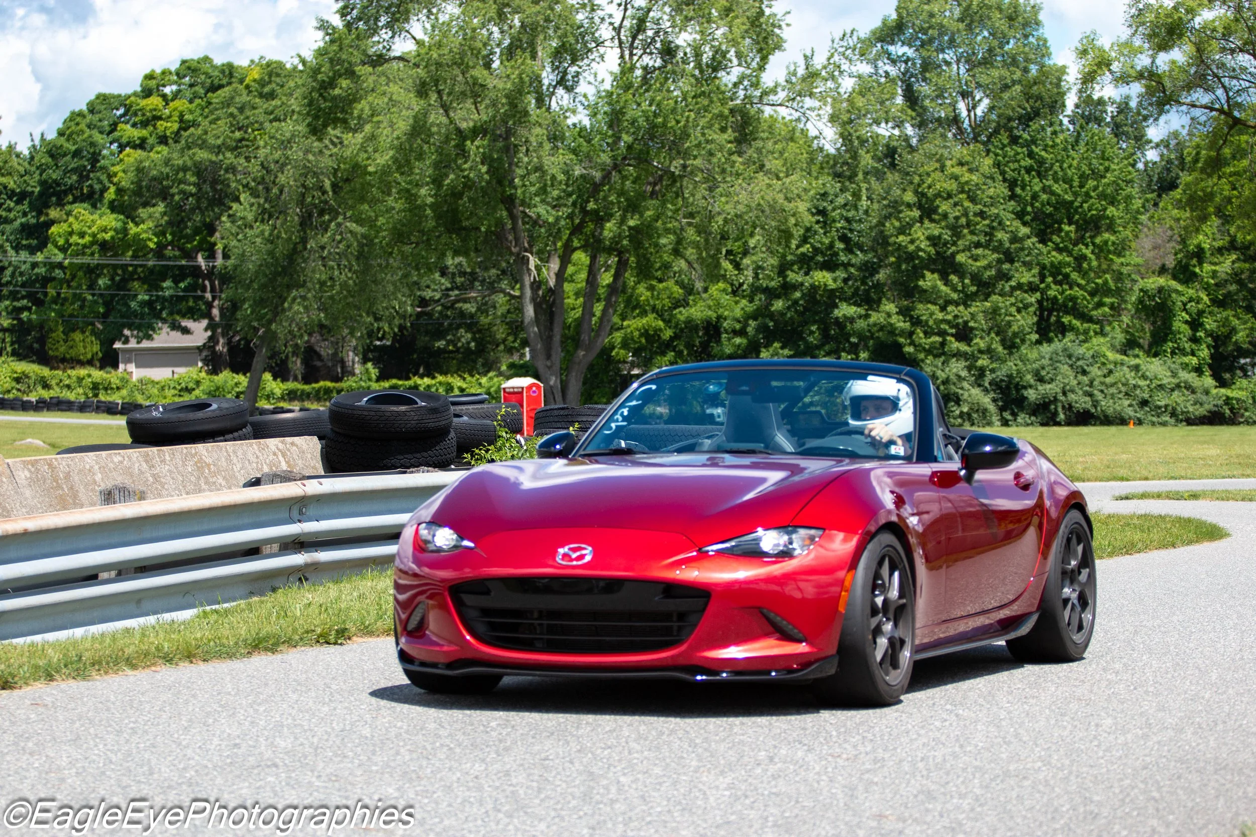 A red Mazda convertible driving on a paved road next to a white safety barrier, with a person wearing a helmet inside. In the background, there are stacks of tires, green trees, and a blue sky with some clouds.