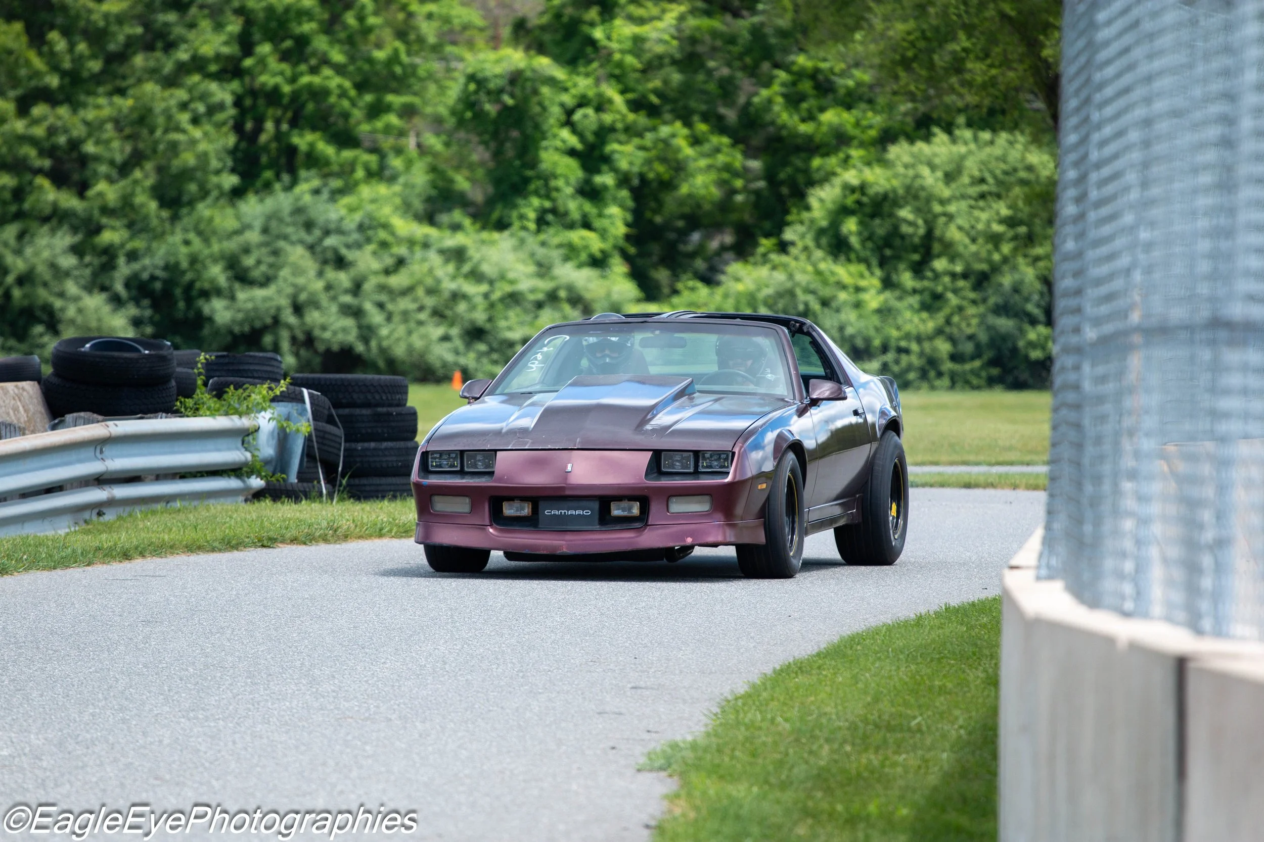 A maroon Chevrolet Camaro sports car driving on a race track surrounded by grass, tires stacked on the side, and lush green trees in the background.