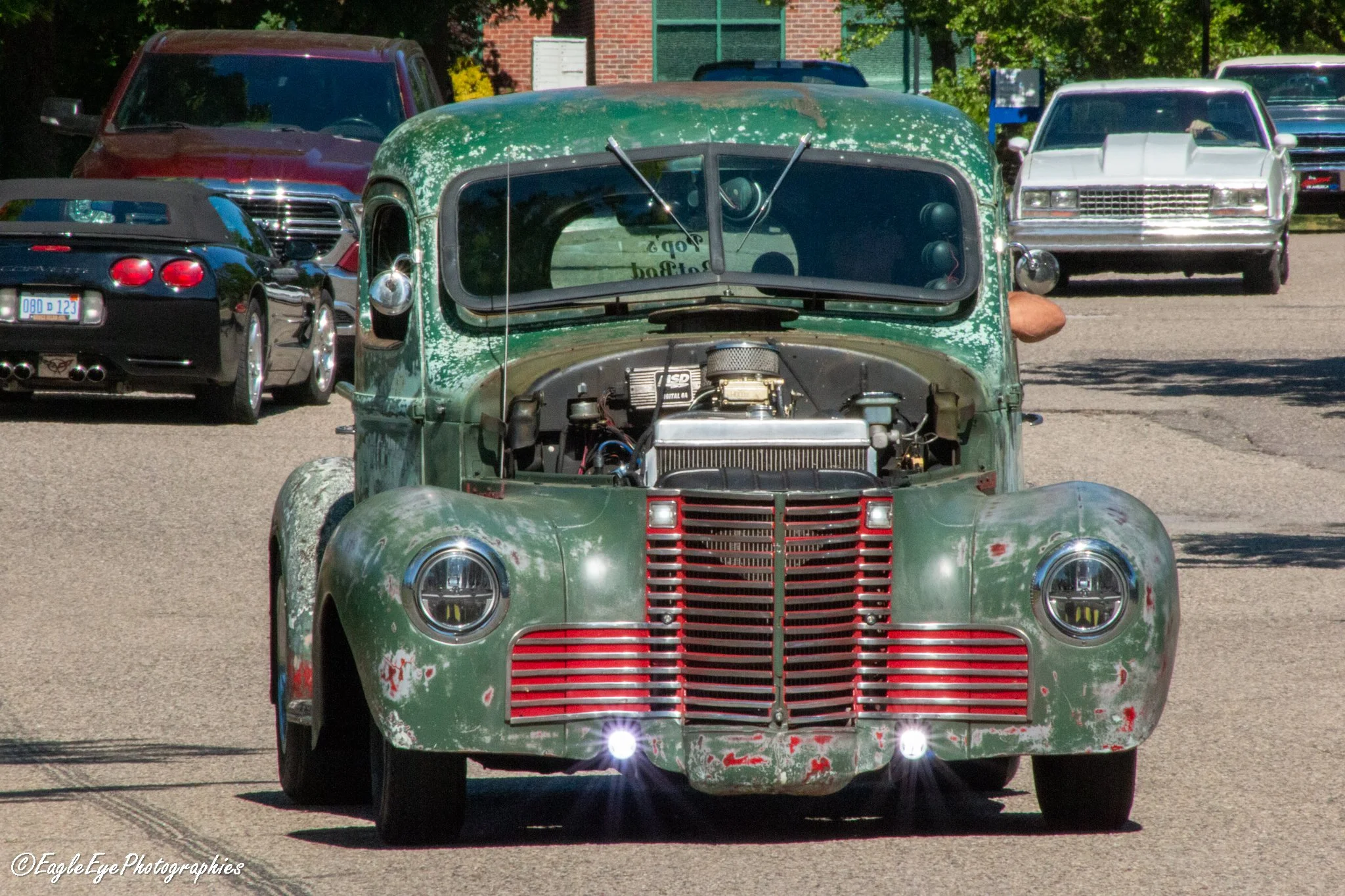 An old, green, rusty pickup truck with its hood open, revealing the engine, parked on a street with other cars and trees in the background.
