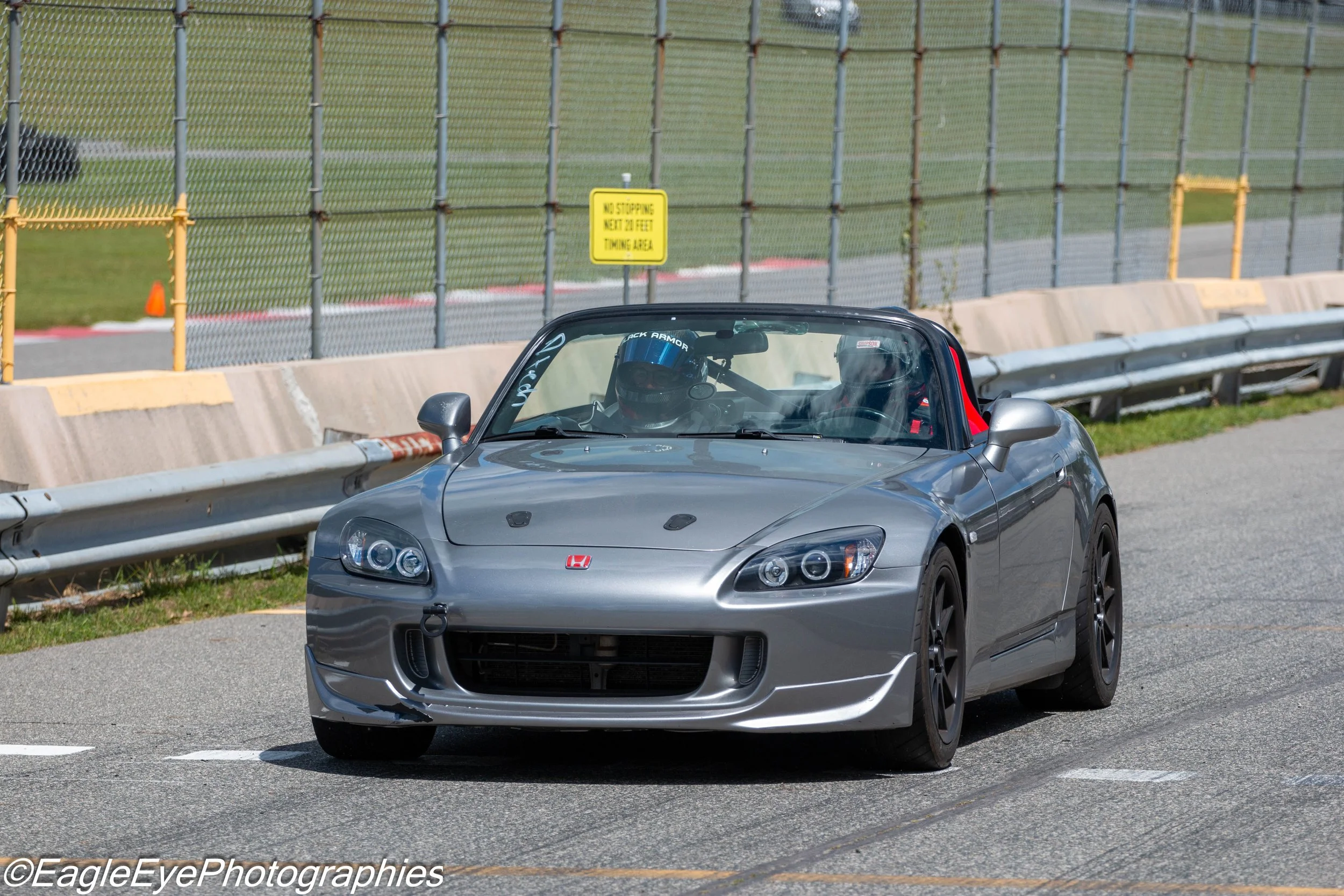 A silver Honda S2000 sports car with a driver wearing a blue helmet is on a racetrack, surrounded by safety barriers and a chain-link fence, with a yellow sign that reads 'NO STOPPING KEEP 20 FEET TOWING AREA' in the background.