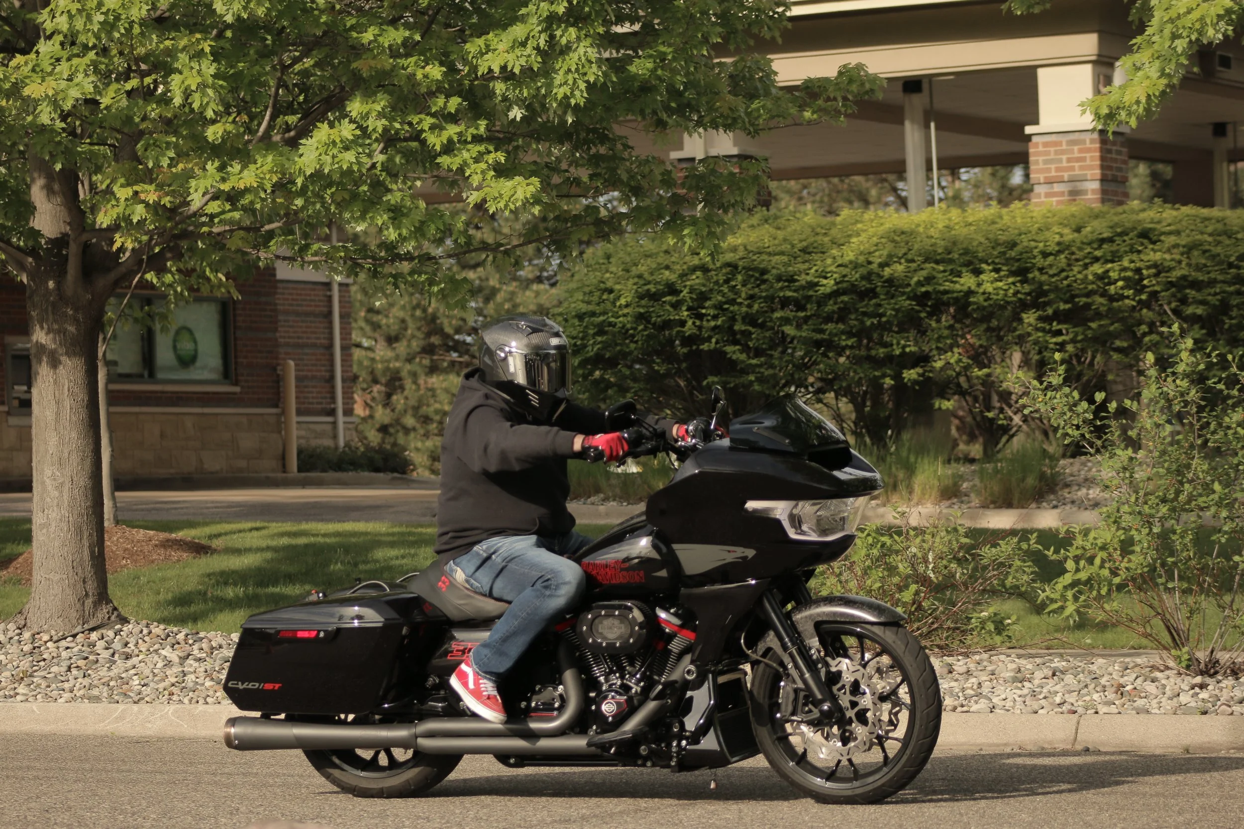 A person wearing a black helmet, black jacket, jeans, and red sneakers riding a black Harley-Davidson motorcycle on a street, with a tree, bushes, and a building in the background.