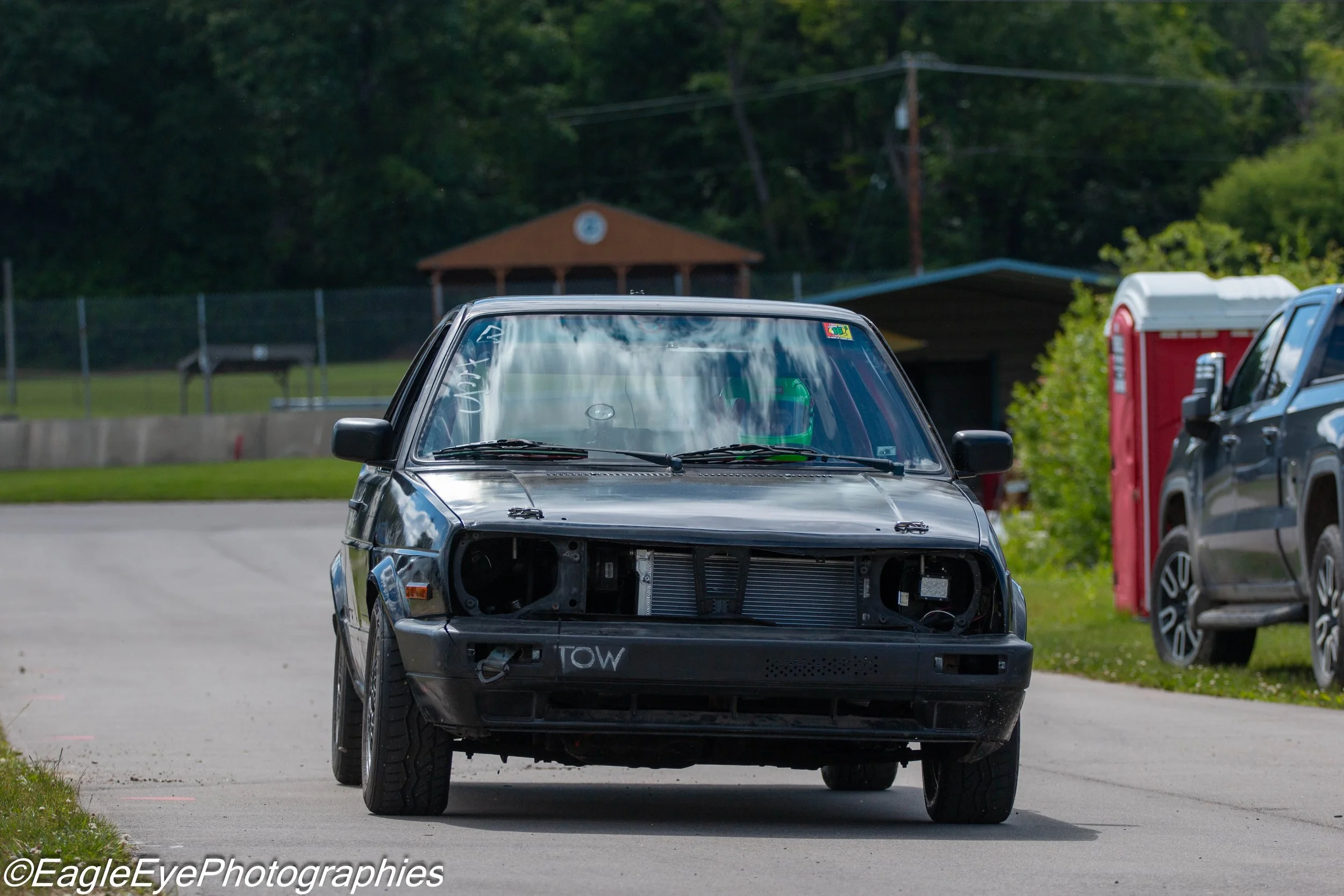 A partly disassembled black car with the front bumper and grille removed, exposing the radiator, parked outdoors near a grassy area, with other vehicles and a red fire hydrant in the background.