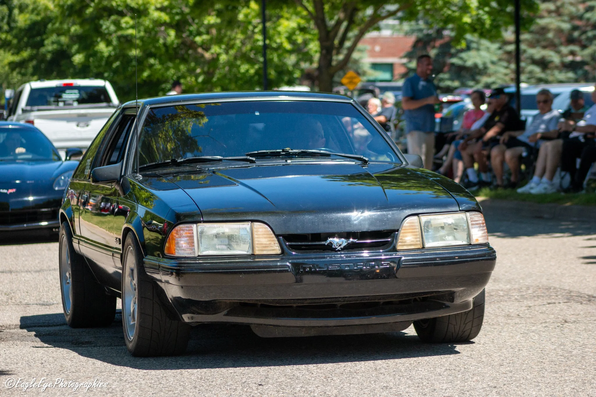 A black vintage Ford Mustang car parked outdoors with a group of people sitting on chairs in the background and green trees around.