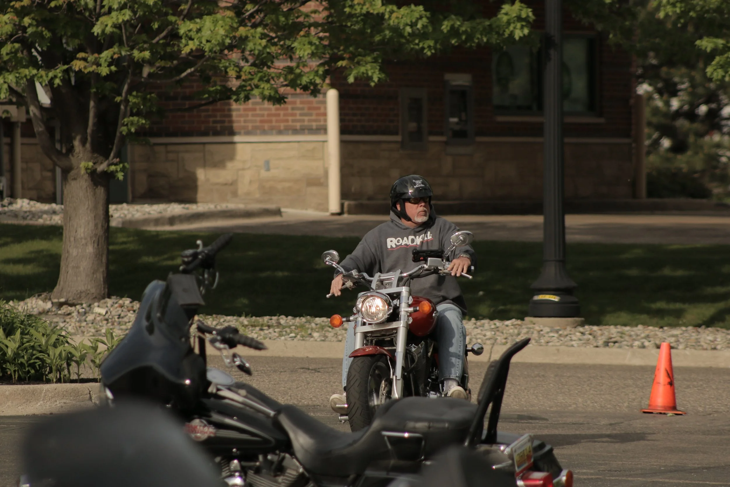 A man wearing a black helmet and sunglasses sitting on a motorcycle, holding its handlebars, with other motorcycles and orange traffic cones nearby in an outdoor setting with trees and a brick building in the background.