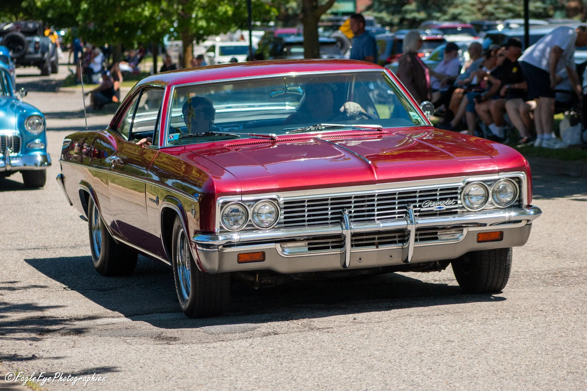 A red vintage Chevrolet car at an outdoor classic car show, with people sitting on benches and walking in the background.