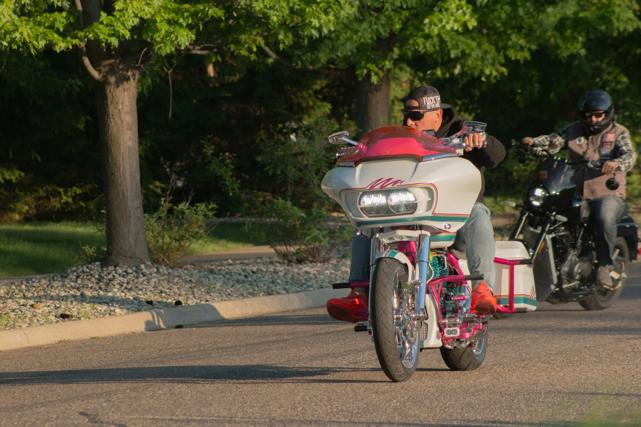 Person riding a pink and white motorcycle with a sidecar on a street, with another motorcyclist behind them, trees in the background.