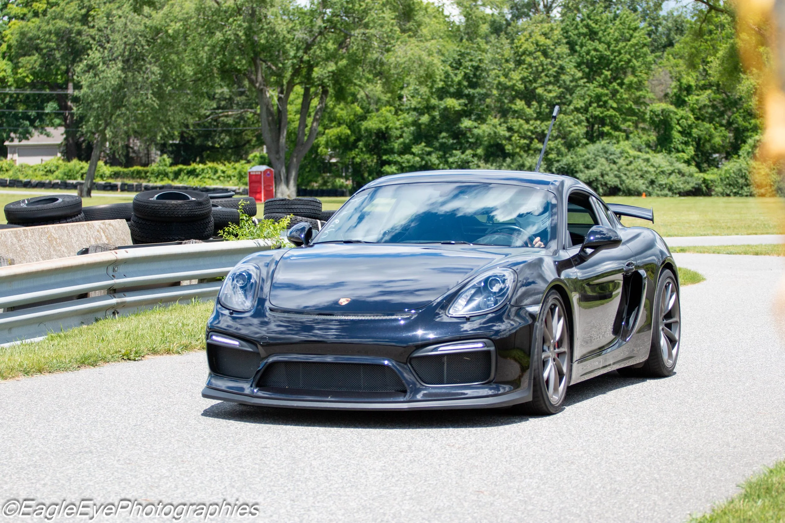 A black Porsche sports car on a paved road, with a line of stacked tires and lush green trees in the background.