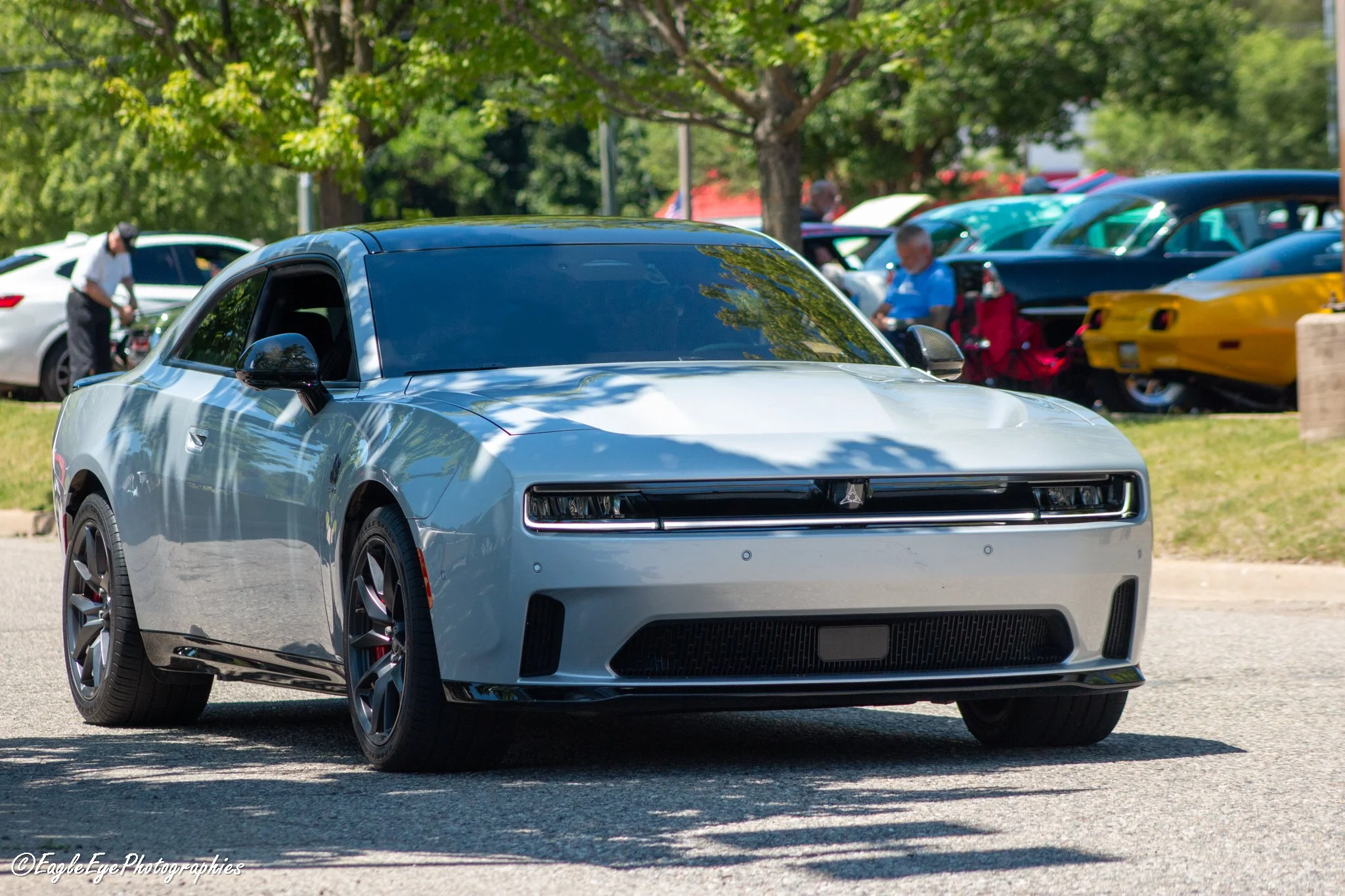 A silver electric sports car parked outdoors at a car show, with trees and other cars in the background, and two people observing the vehicles.