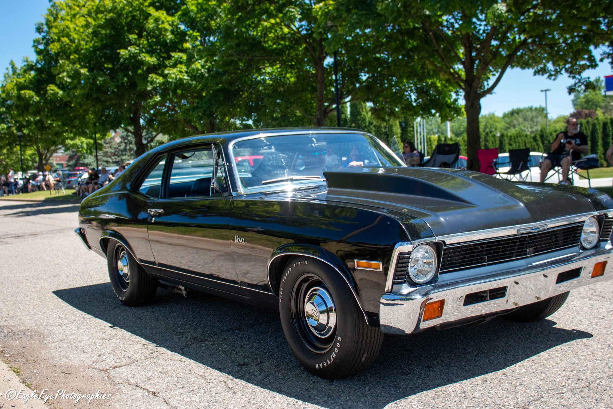 Black classic Chevrolet Monte Carlo car parked at an outdoor event with people sitting in chairs under leafy trees.