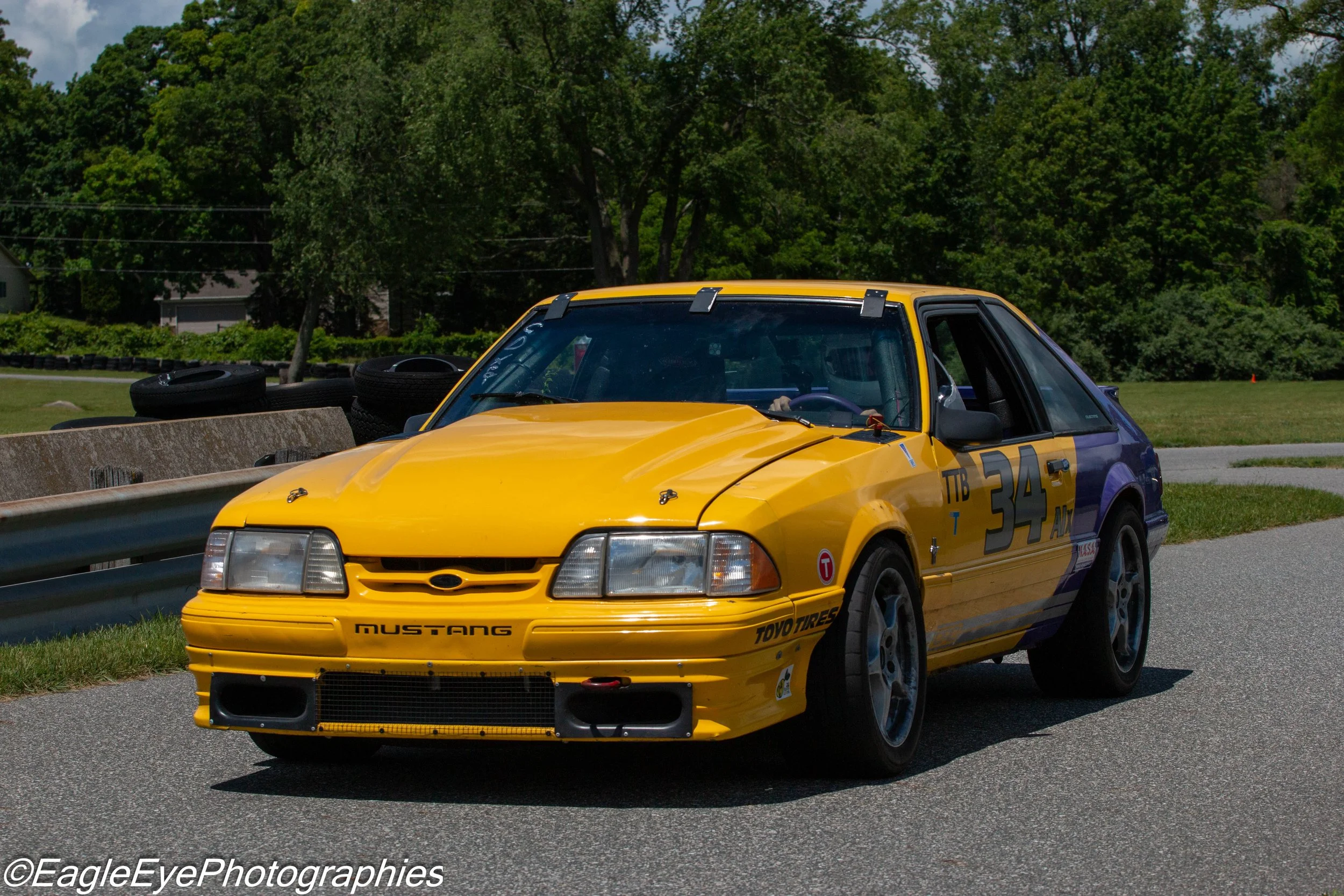 A yellow race car with racing decals and the words 'Mustang' and 'Toyo Tires' on the front, driving on a road near a barrier, with green trees and a cloudy sky in the background.