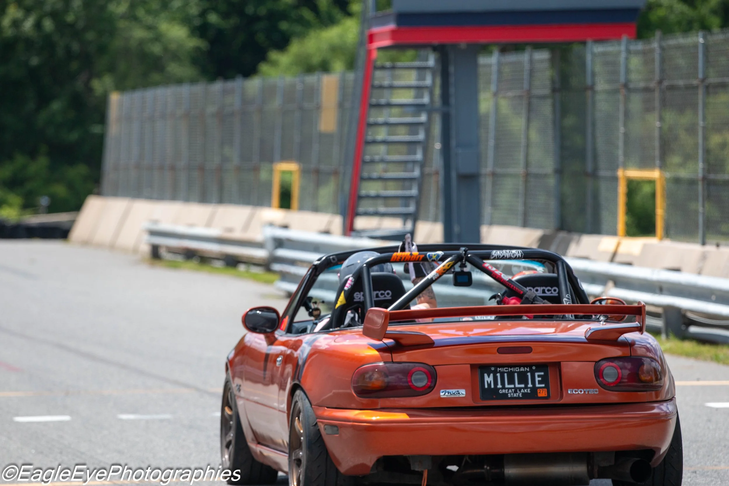A red convertible toy car with a Michigan license plate reading 'MILLIE' and the name tag 'Great Lake State' on the rear, parked on the side of a road. The car has a roll cage, racing seats, and visible helmets inside. In the background, there is a t