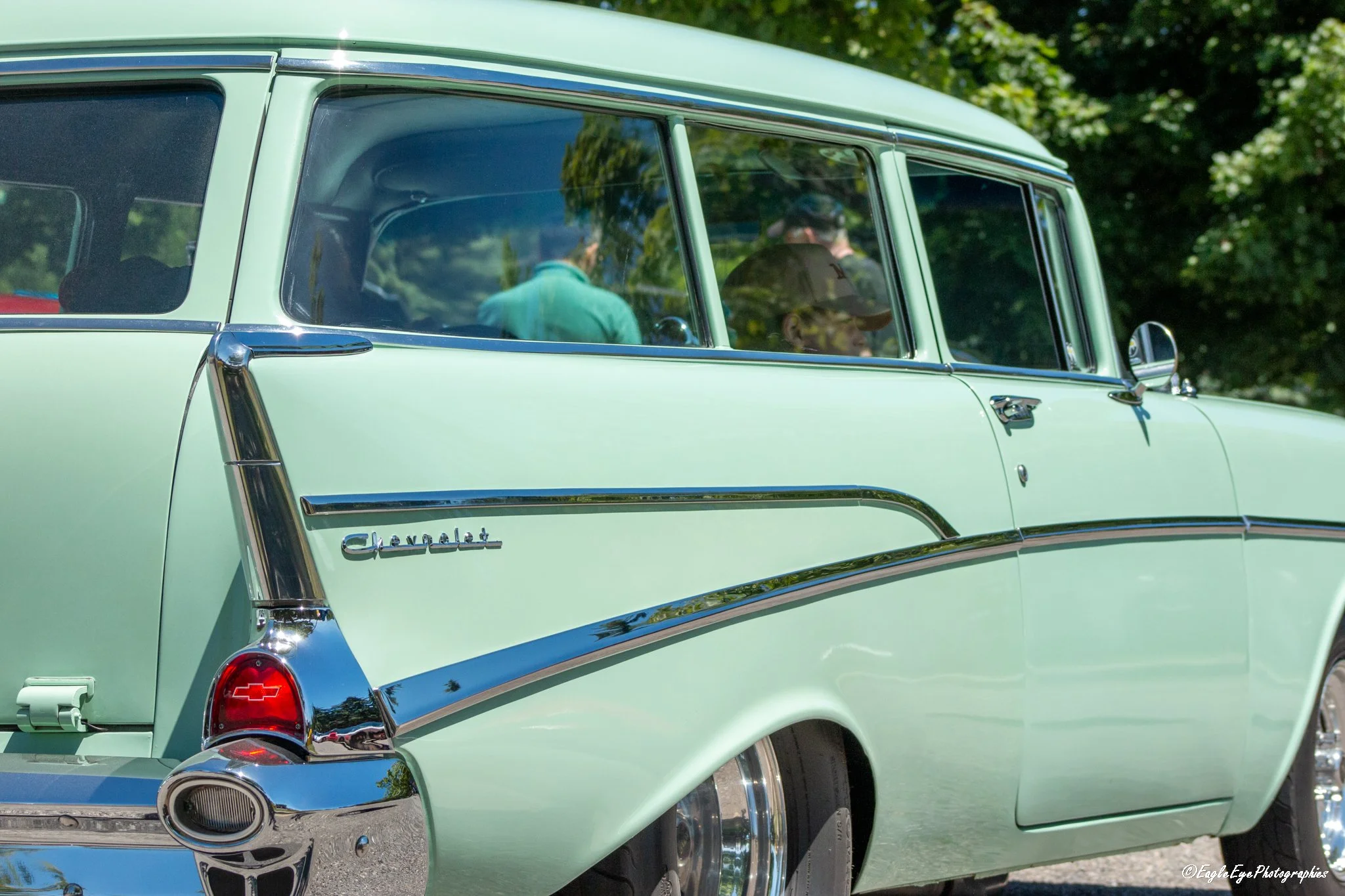 A vintage light green Chevrolet Suburban with chrome details, parked outdoors with a background of trees and greenery. The image captures the mid-section of the vehicle, showing the Chevrolet emblem and rear tail light.