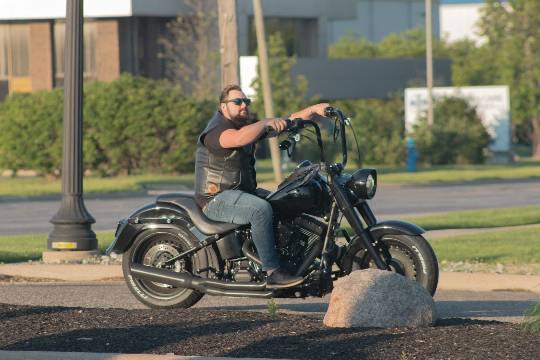 A man with a beard and sunglasses riding a black motorcycle on a city street with green bushes and buildings in the background.