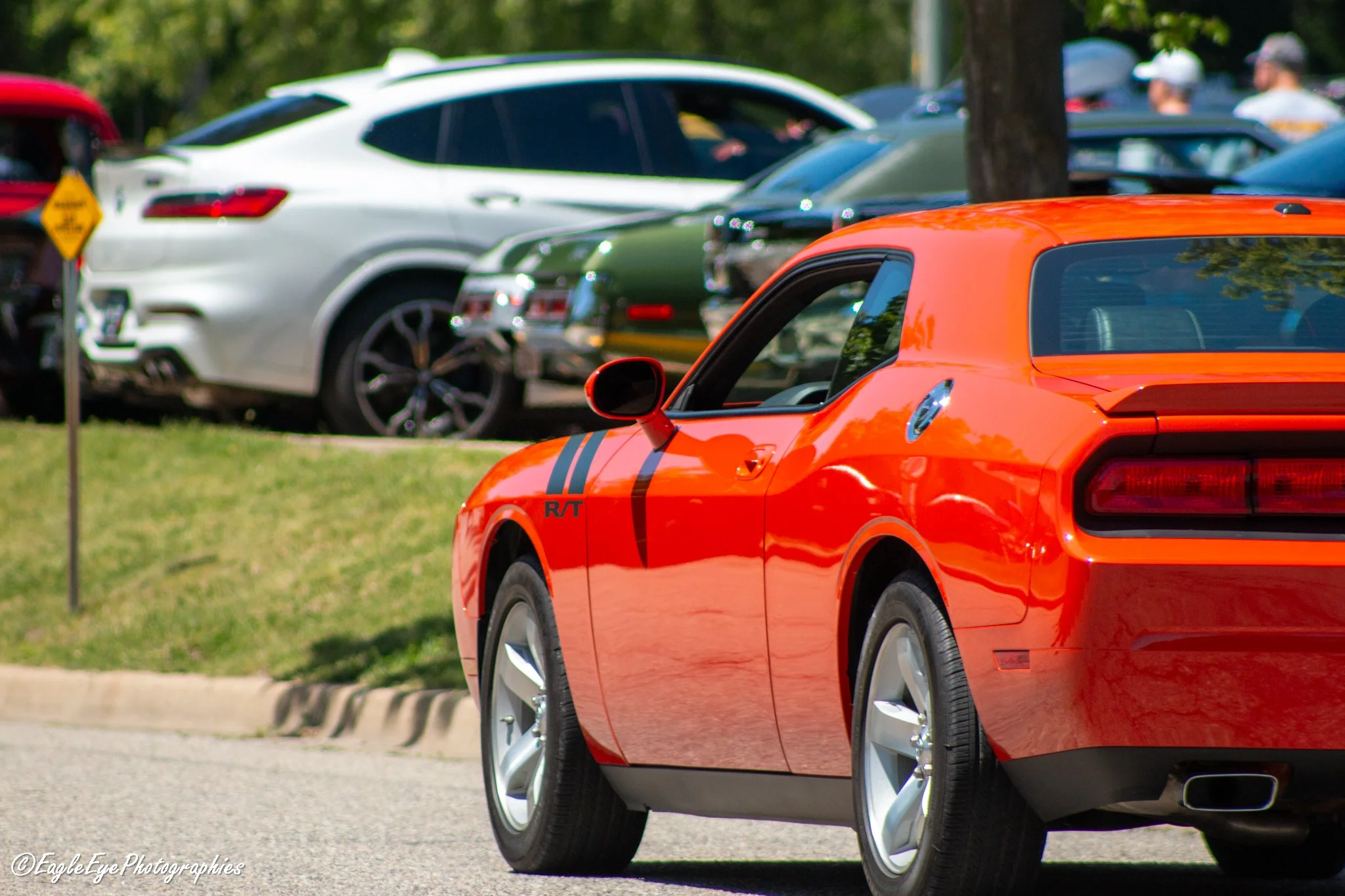 Close-up of an orange sports car, likely a Dodge Challenger, parked in a lot with other cars and trees in the background.