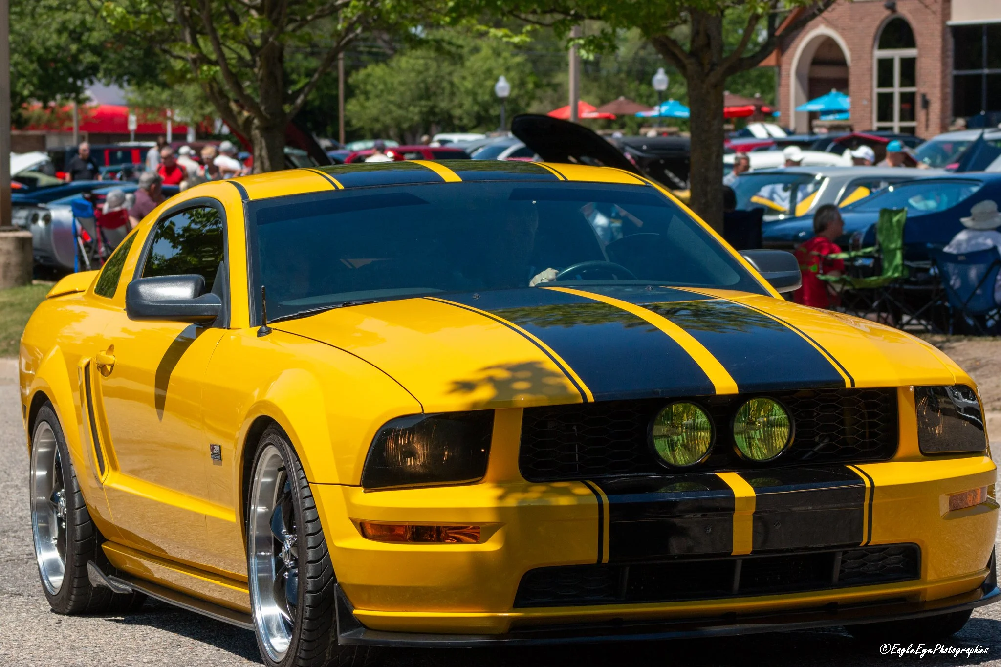 Yellow sports car with black racing stripes displayed at an outdoor car show, with people walking and sitting in the background under trees.