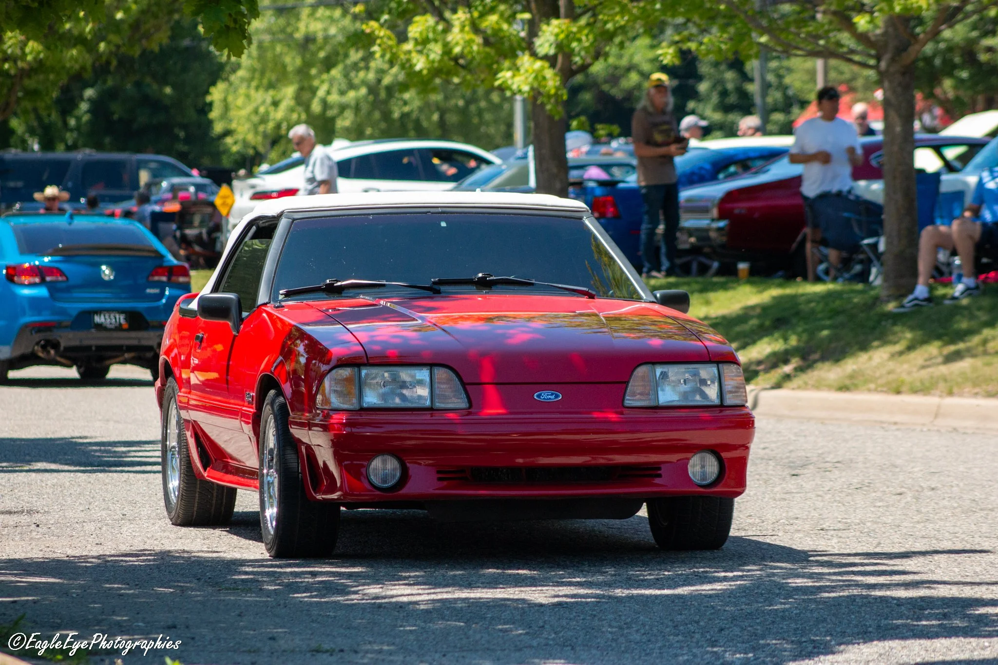 Red sports car parked in front of a crowd of people at an outdoor event with trees in the background.